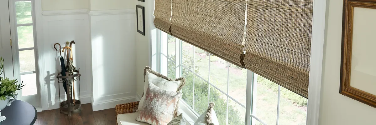 Interior of a cozy room with a wicker armchair, decorative pillows, large windows with woven bamboo blinds, and an umbrella stand with umbrellas in the corner.