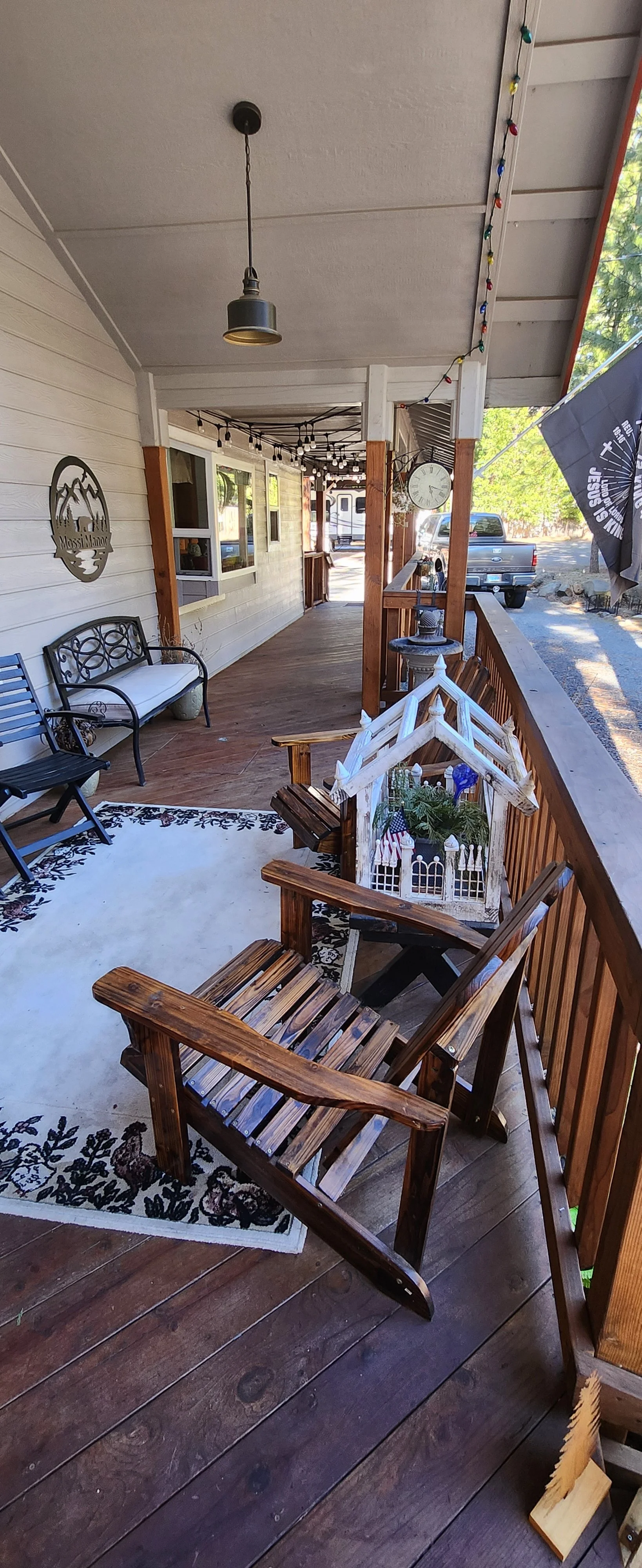A porch with a wooden floor and railing. There are outdoor chairs, a bench, a small table with decorations, string lights, a clock on the wall, and a decorative sign with mountains and the words "Mossy Mountain."
