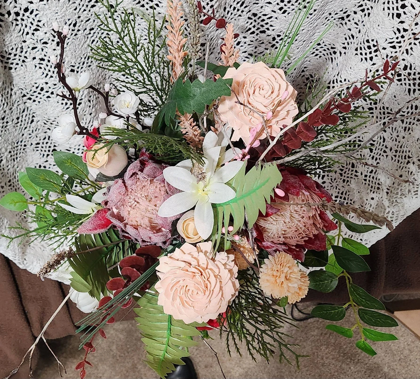 A bouquet of artificial flowers with pale peach roses, white orchids, greenery, and other plant stems and leaves, placed on a lace tablecloth.