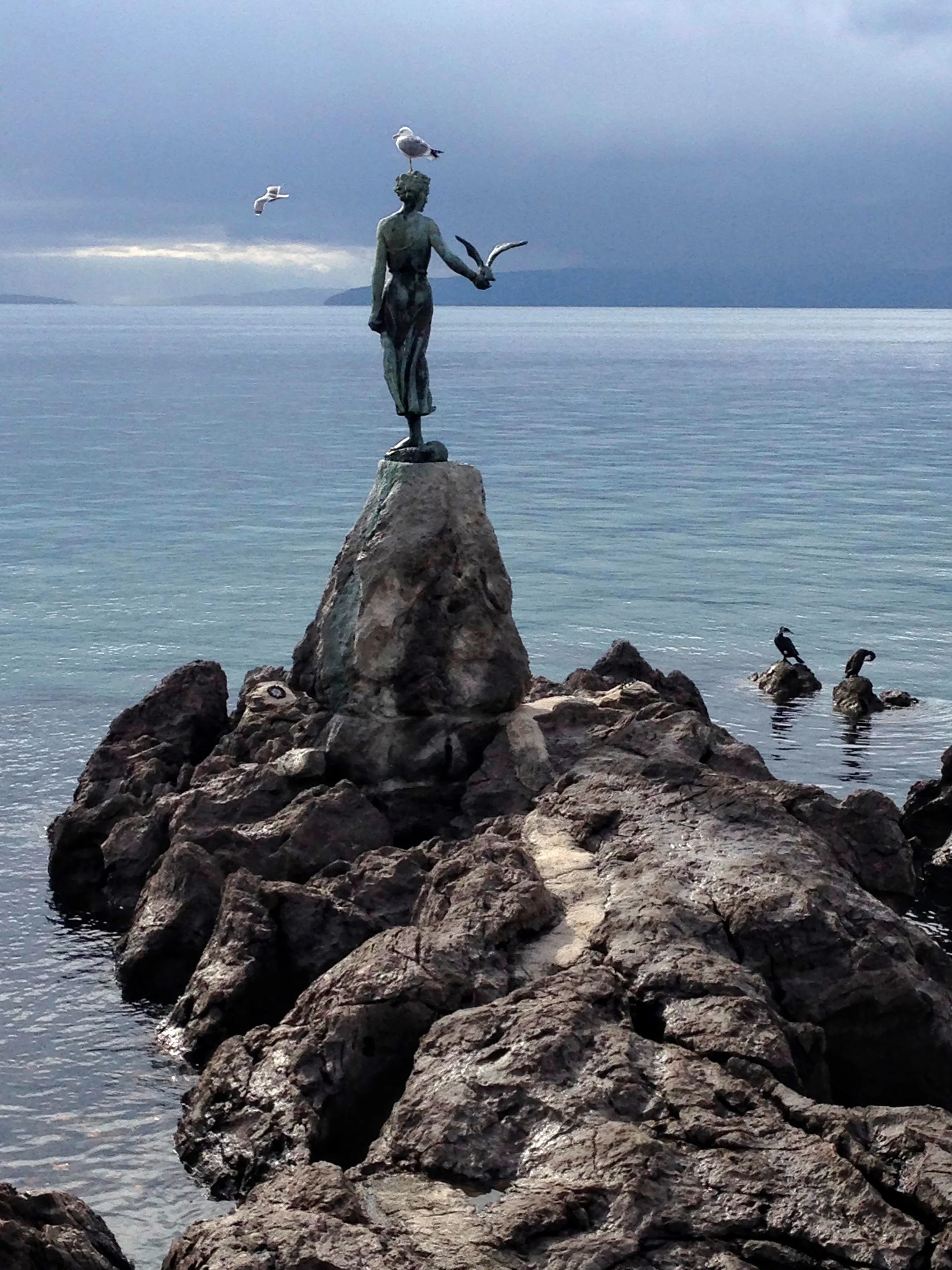 Sculpture of a woman with a bird appearing at the edge of rocks on the ocean with real seagulls at play