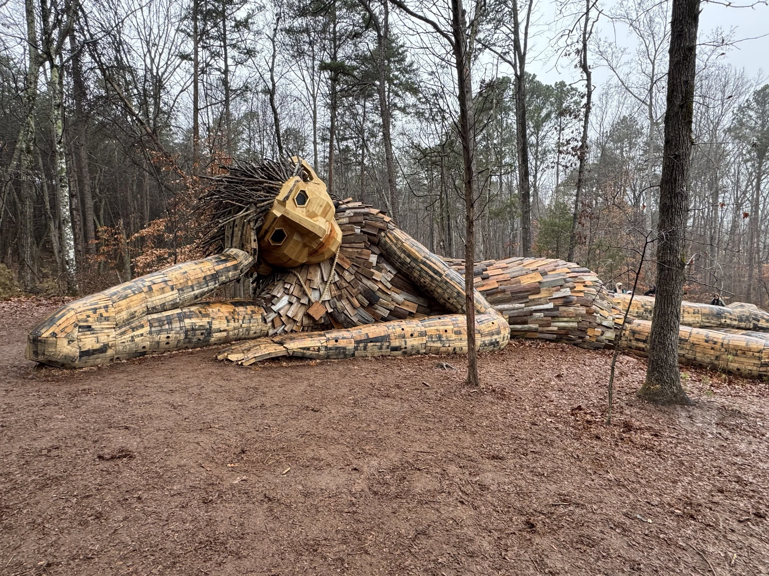 A large wooden sculpture of a reclining man in a forest clearing, surrounded by trees with mostly bare branches.