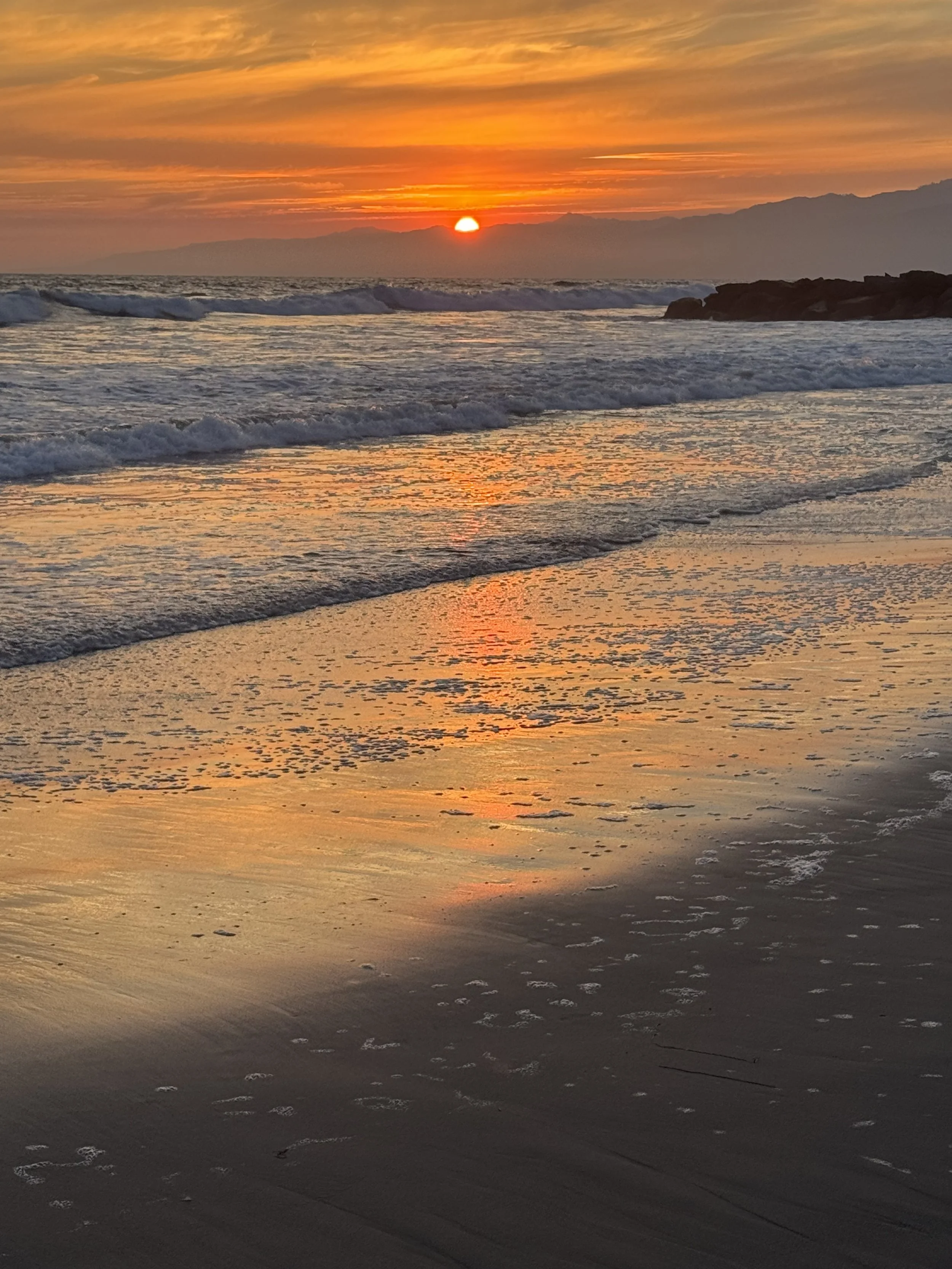 Sunset over the ocean with waves crashing on the shore and a rocky outcrop on the right side, with colorful orange and yellow clouds in the sky.