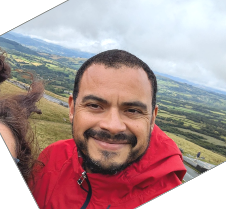 A man with short dark hair and a beard, smiling, outdoors with a scenic landscape of rolling hills and cloudy sky in the background.