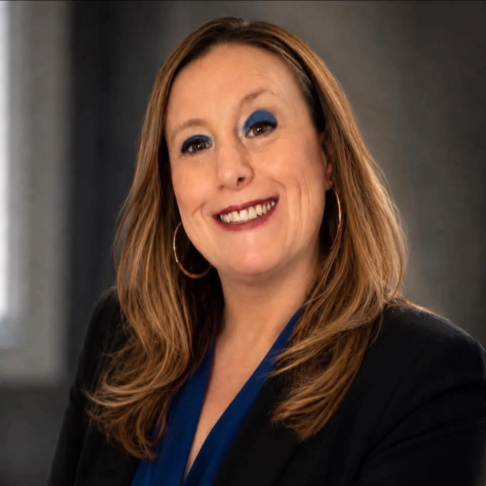 Portrait of a woman with long reddish-brown hair, wearing blue eyeshadow, hoop earrings, a black blazer, and a blue top, smiling at the camera.