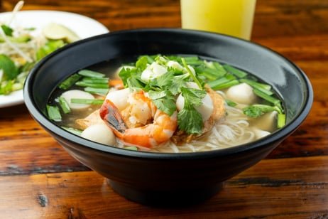 A bowl of shrimp and vegetable soup garnished with cilantro, served on a wooden table with a side salad and a glass of yellow beverage in the background.