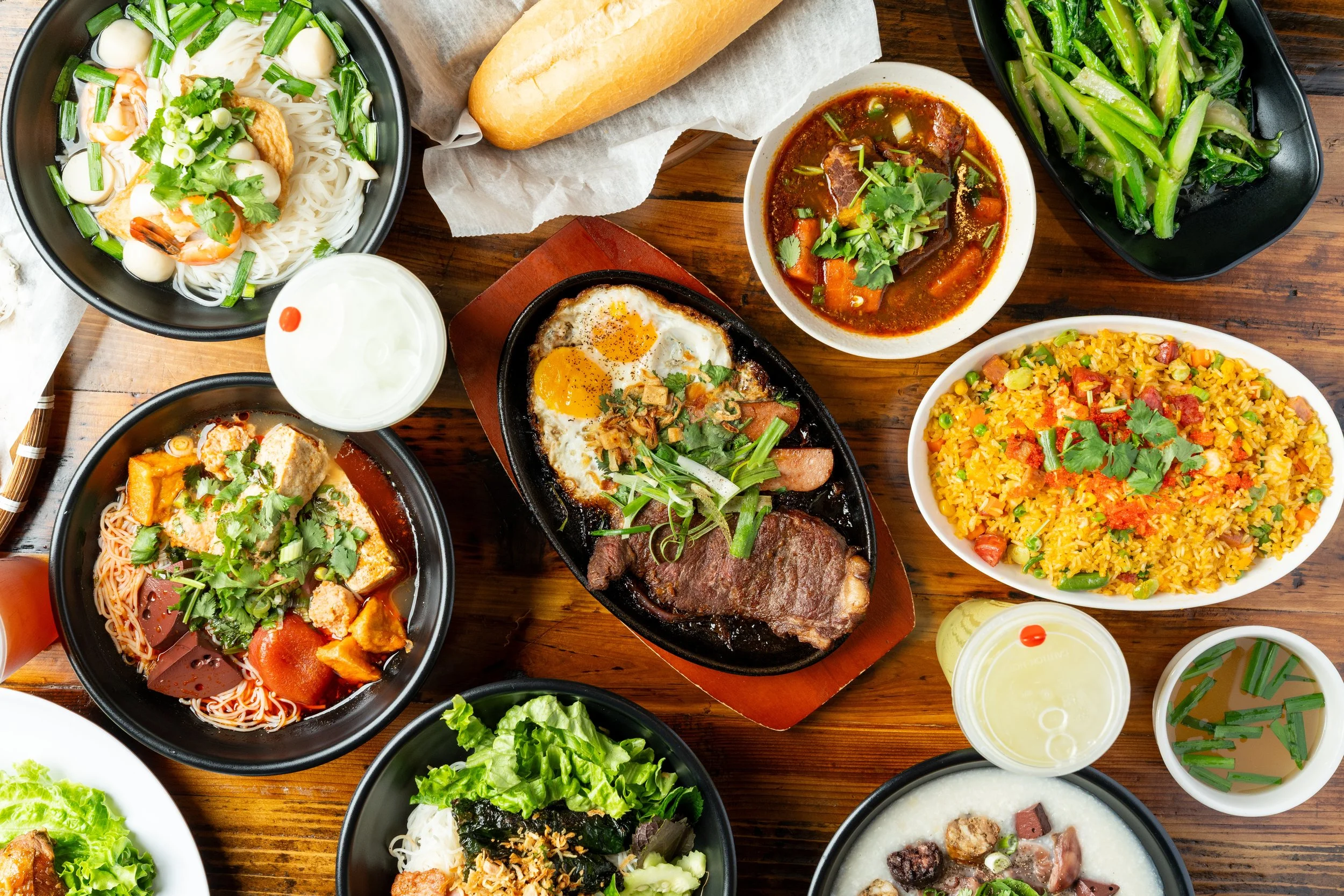 A variety of Asian dishes on a wooden table, including noodle soups, fried rice, a beef stew, a sunny side up egg, and fresh green vegetables.