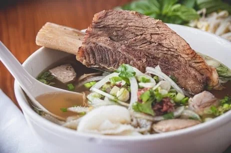 A bowl of Vietnamese pho with a large piece of beef, herbs, and noodles, with a spoon inside.