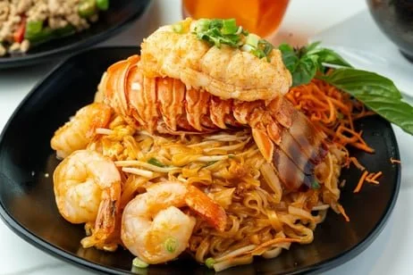 Seafood dish with shrimp, fried fish fillet, noodles, vegetables, and leafy greens on a black plate.