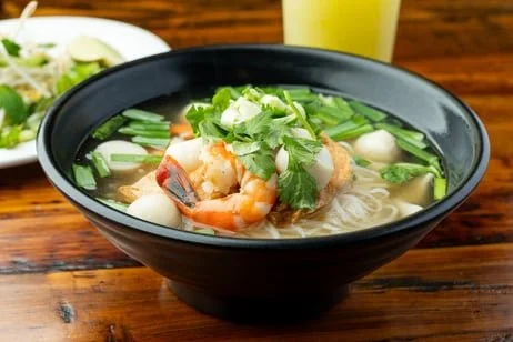 A bowl of Vietnamese pho with shrimp, white noodles, green onions, cilantro, and seafood, on a wooden table with a side salad and a glass of yellow beverage.