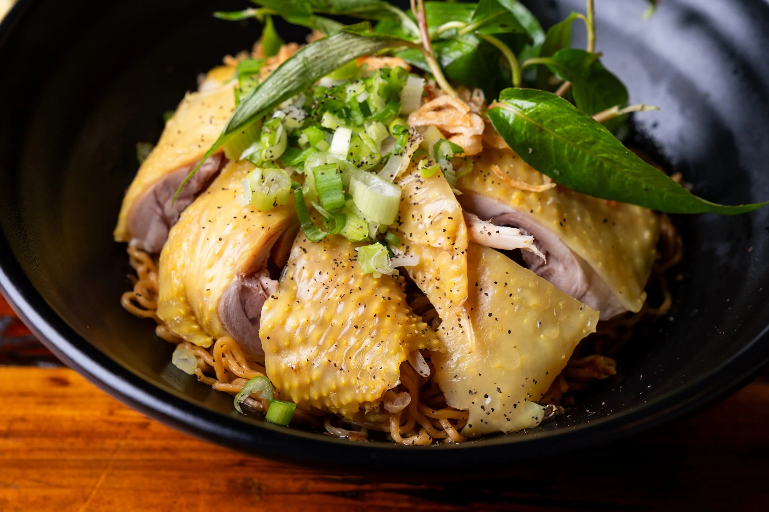 Close-up of a bowl of ramen topped with slices of cooked chicken, chopped green onions, fresh green herb leaves, and black pepper, on a wooden surface.