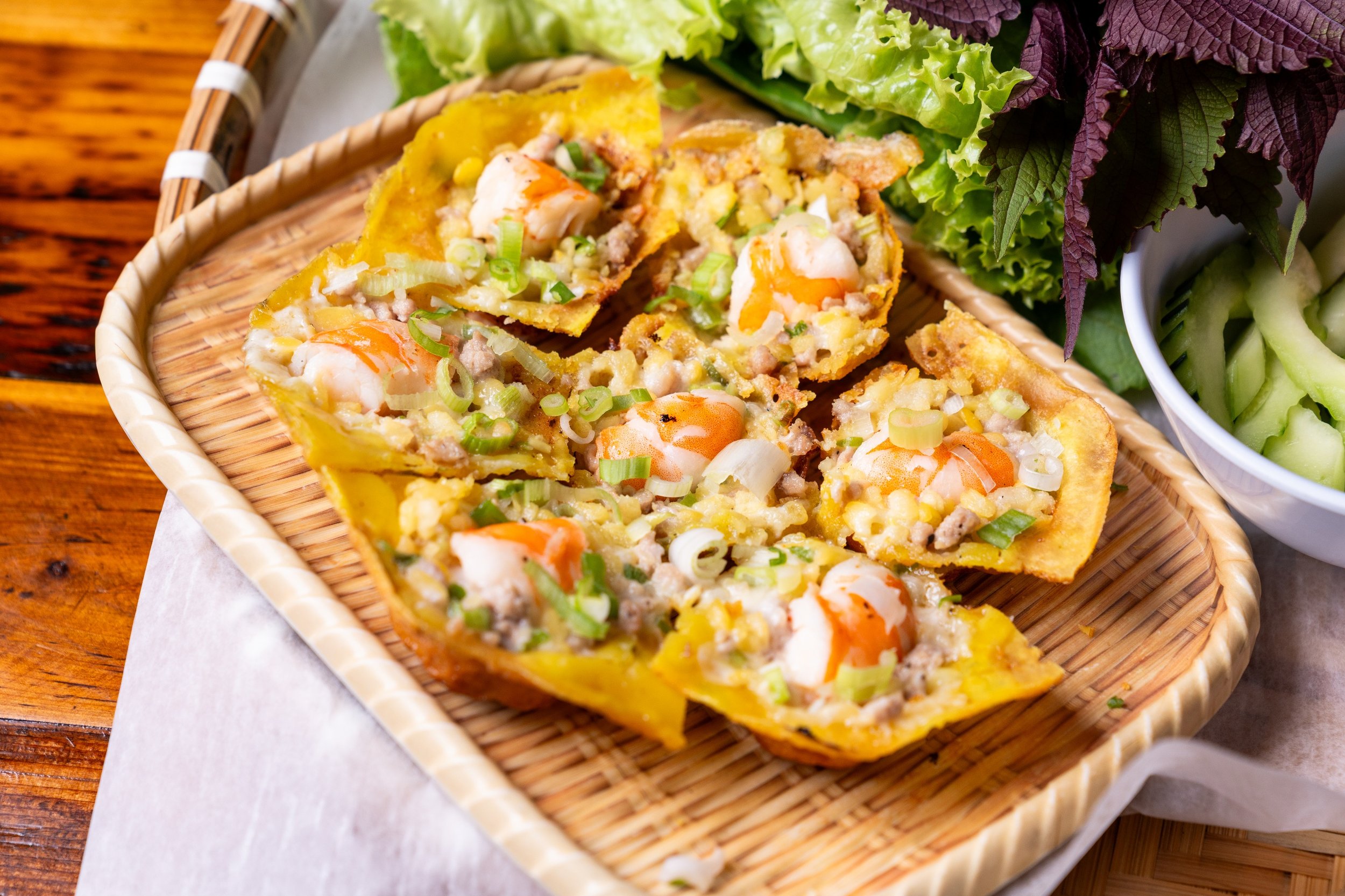 Close-up of a basket with sliced egg foo young, topped with chopped green onions, served on a white paper, with fresh leafy greens in the background on a wooden surface.