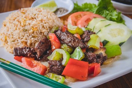 A plate of cooked beef stir-fry with green bell peppers and red peppers, a serving of rice, and fresh sliced cucumbers and tomatoes with lettuce and a lime wedge in the background.