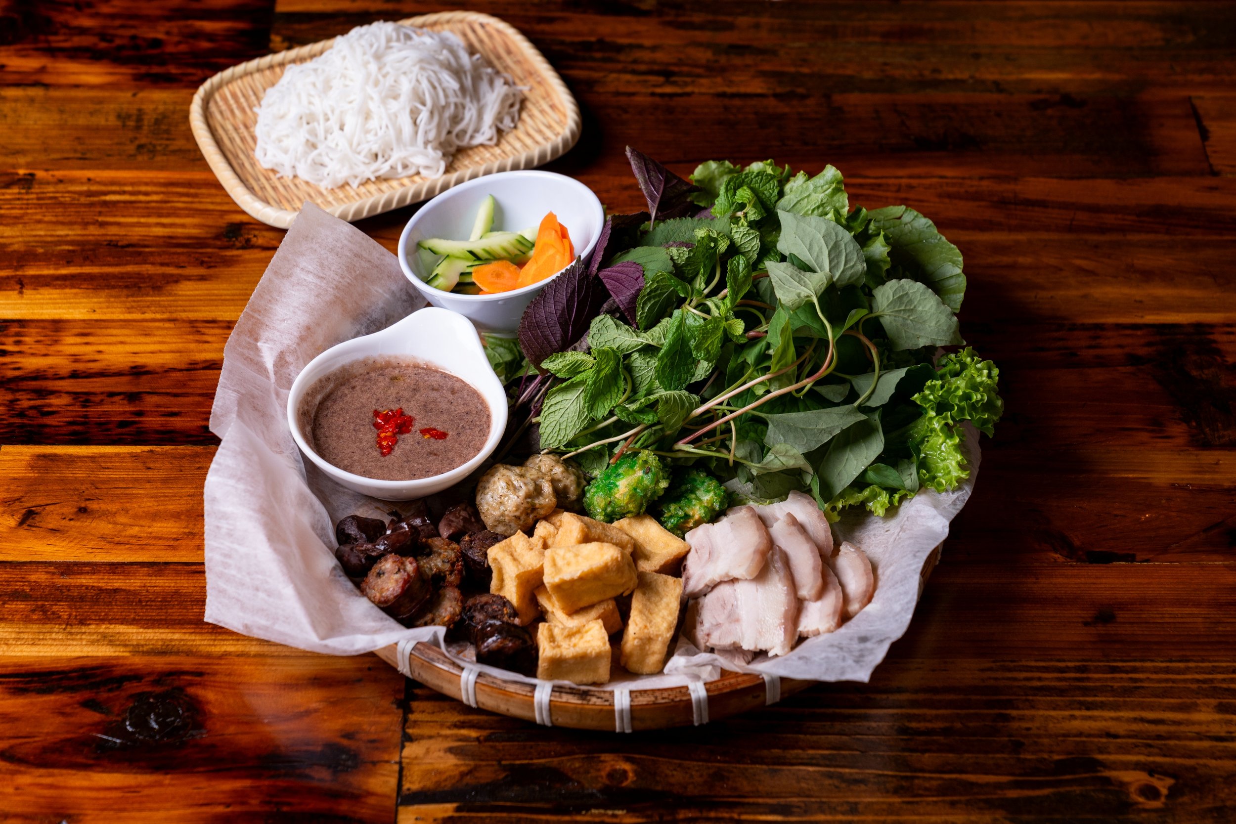 Assorted Vietnamese dishes on a round bamboo tray, including rice noodles, fresh herbs, dipping sauces, and various grilled and fried meats on a wooden table.