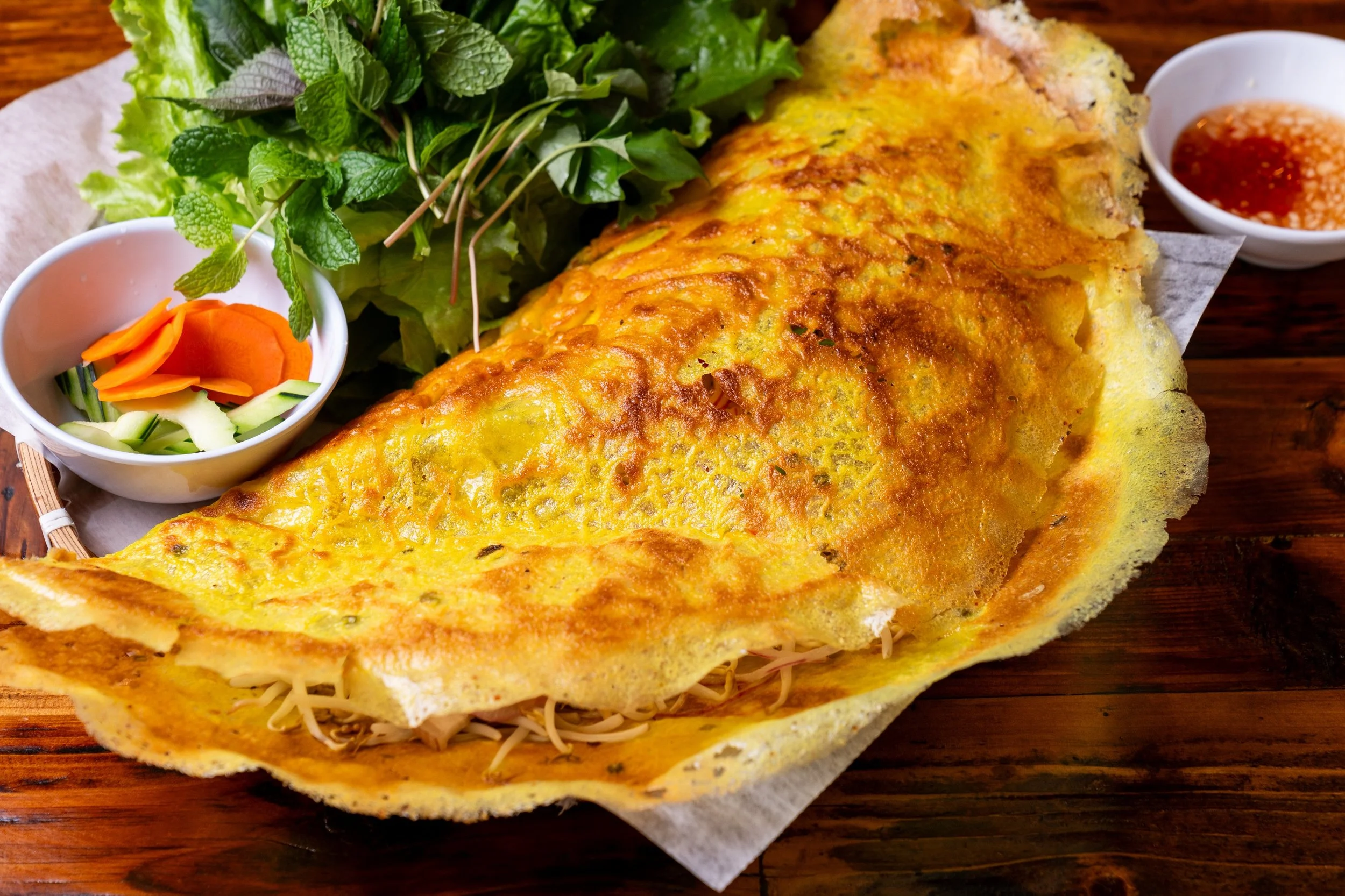 Omelet served on a wooden table with a fresh green salad, sliced vegetables, and two small bowls of dipping sauce.