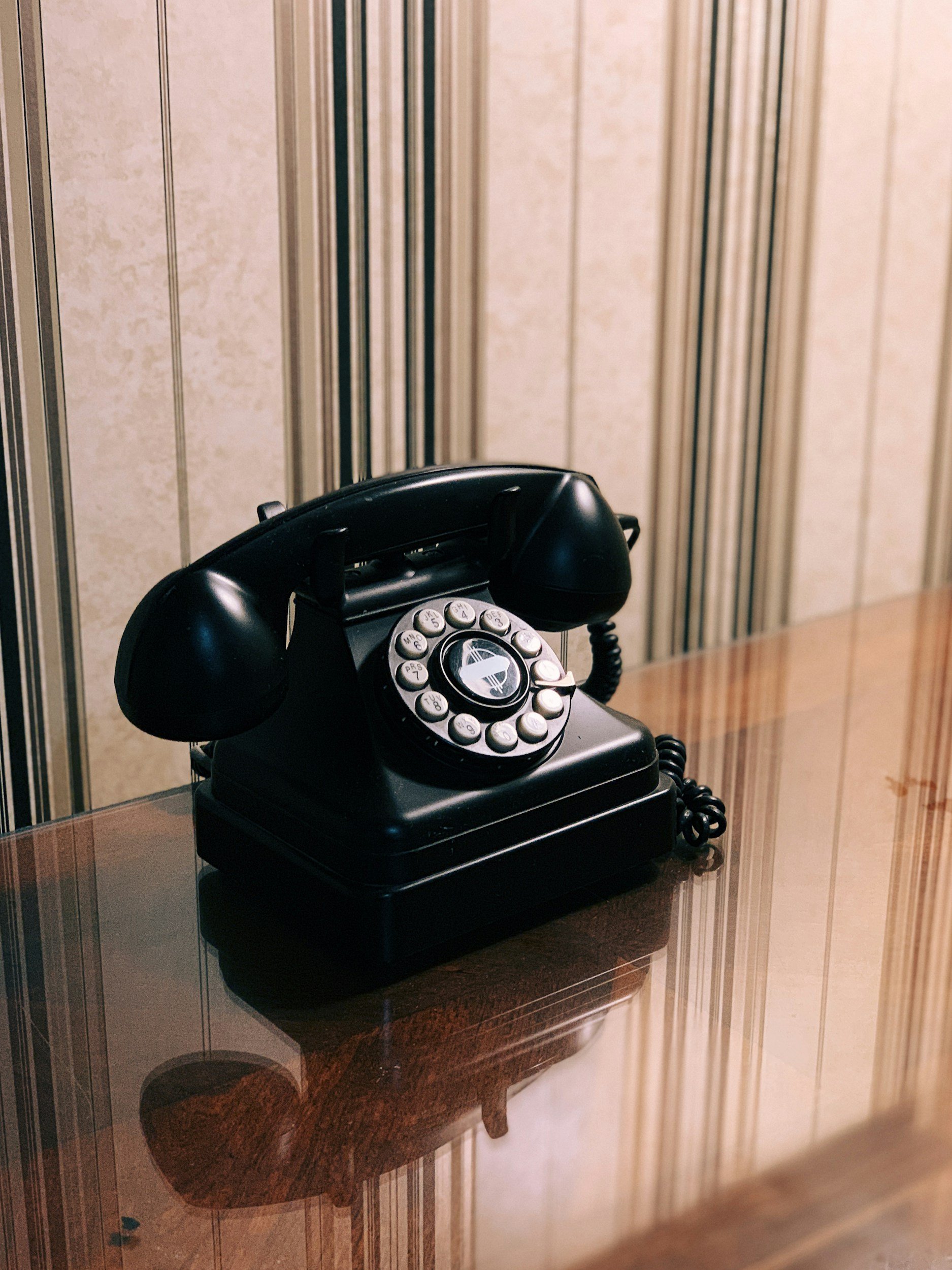 A black vintage rotary telephone sitting on a polished wooden table with a marbled wall in the background.