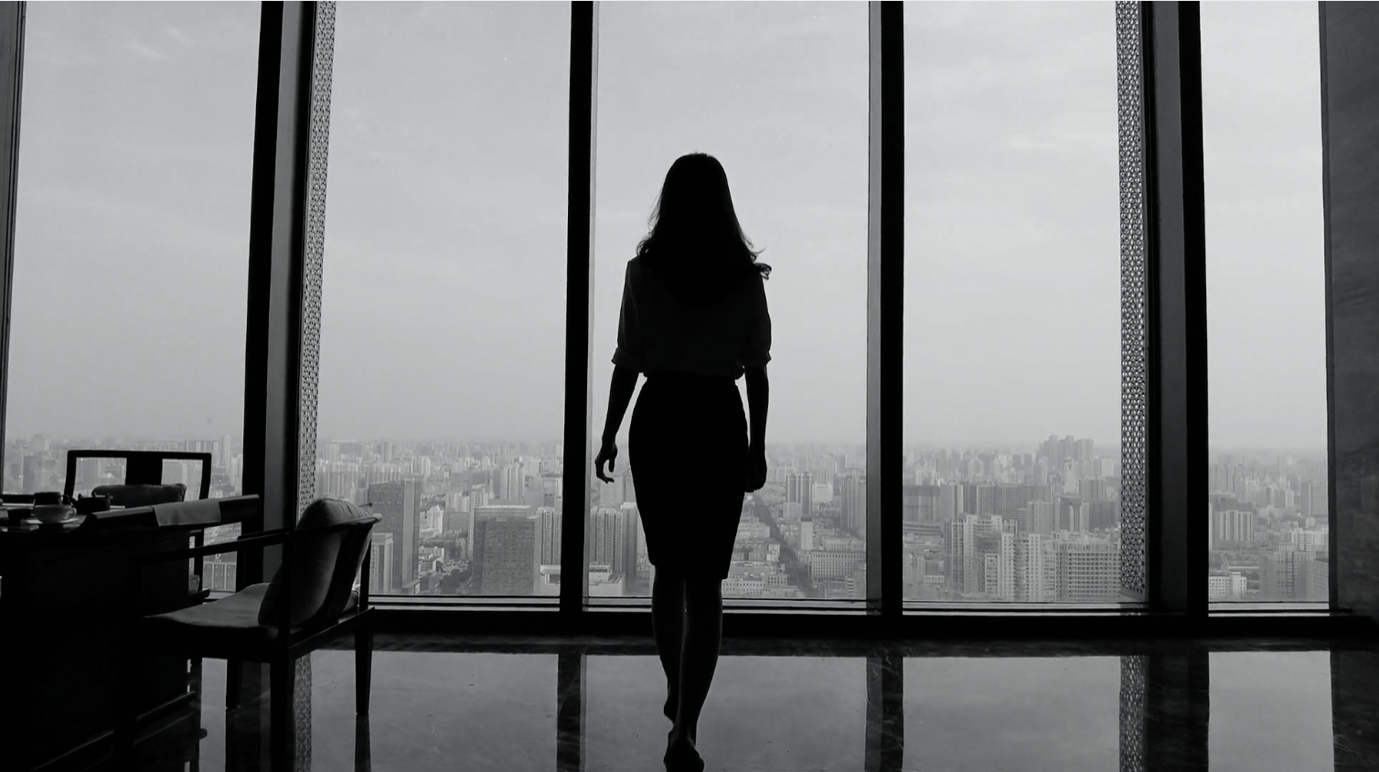 A silhouette of a woman standing inside a high-rise building with large floor-to-ceiling windows, overlooking a city skyline in the background.
