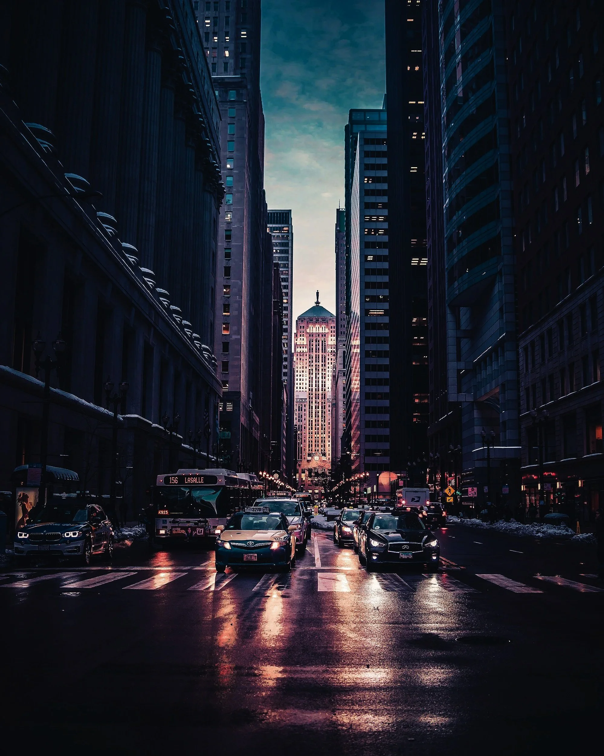 Dark city street at dusk with wet pavement reflecting city lights, tall skyscrapers on either side, and a bus and cars at a stop.