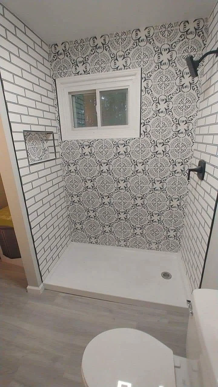 A shower area with patterned black and white tiles on the back wall, white brick-patterned tiles on the side walls, a small window, and a silver drain in the floor. Part of a toilet is visible in the foreground.