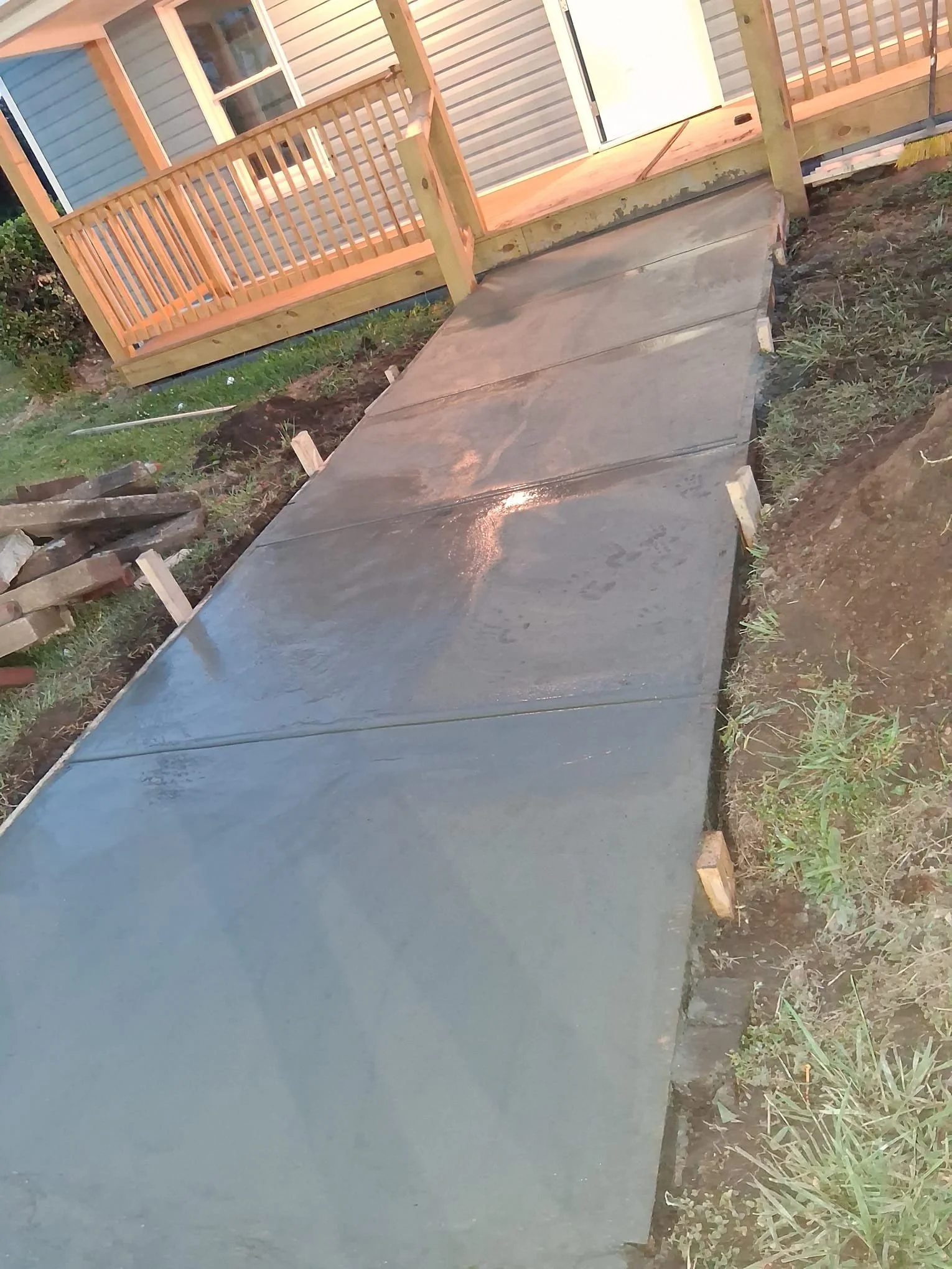 Concrete pathway leading to a house with a small wooden ramp and railing, surrounded by grass and dirt.