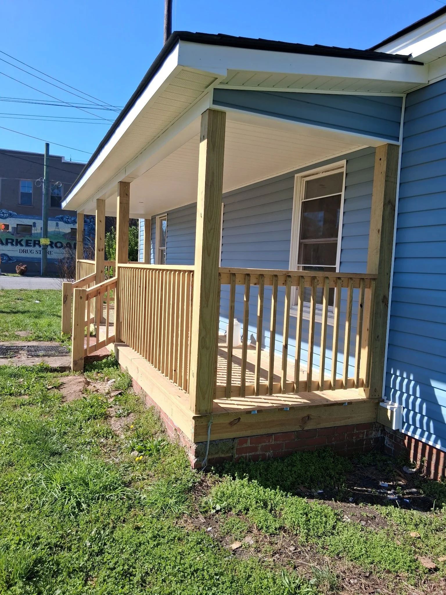 A newly built small porch with a wooden railing and support posts attached to a light blue house. The porch is on a brick foundation, and the grass in the yard is green. In the background, there is a building with a sign partially visible and a clear blue sky.