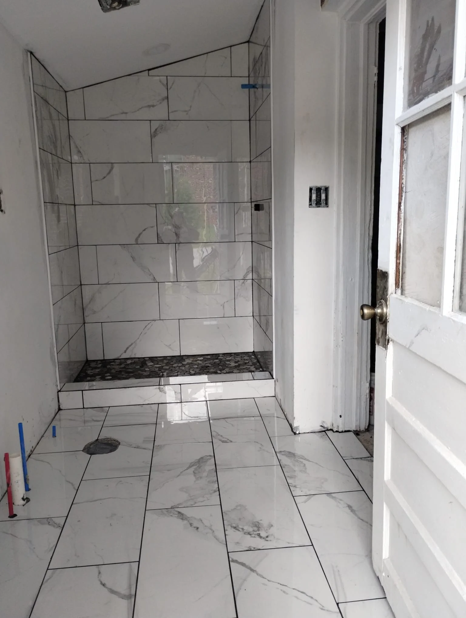 A bathroom under construction with white marble-look tiles on the walls and floor. The shower area features a pebble tile border and the shower drain is visible. There is an open door with a brass doorknob to the right, and unfinished electrical outlets and exposed pipes are seen.