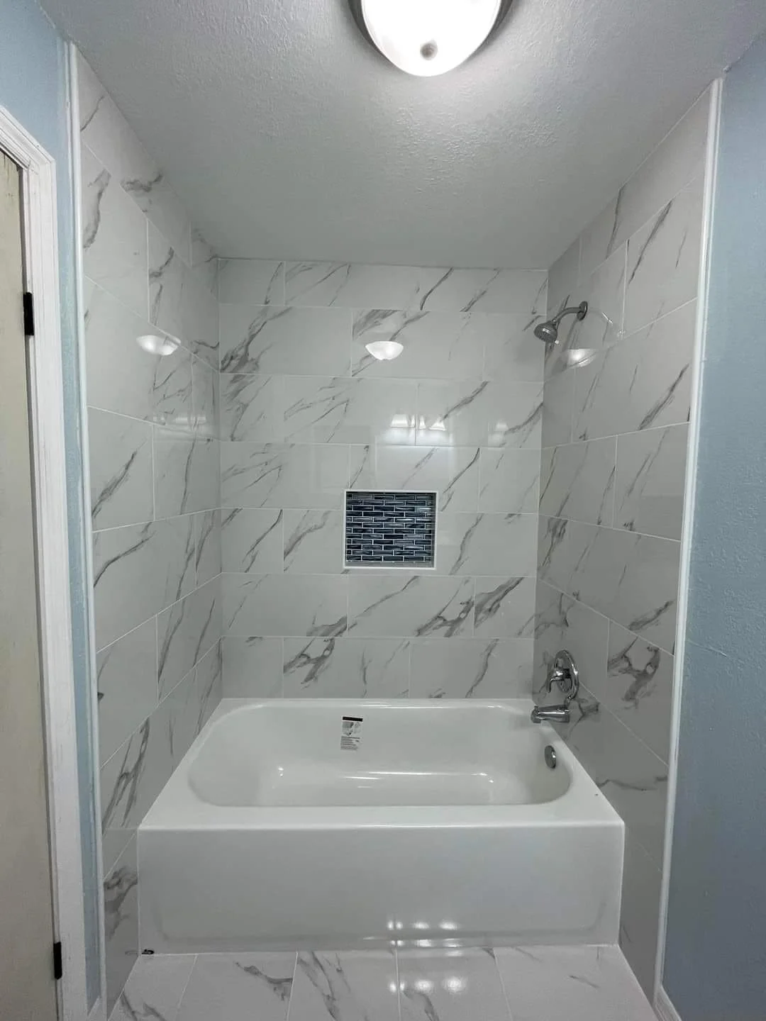 A bathroom shower with marble-patterned tiles, a bathtub, a ceiling light, a showerhead, and a small vent or niche with dark tile.