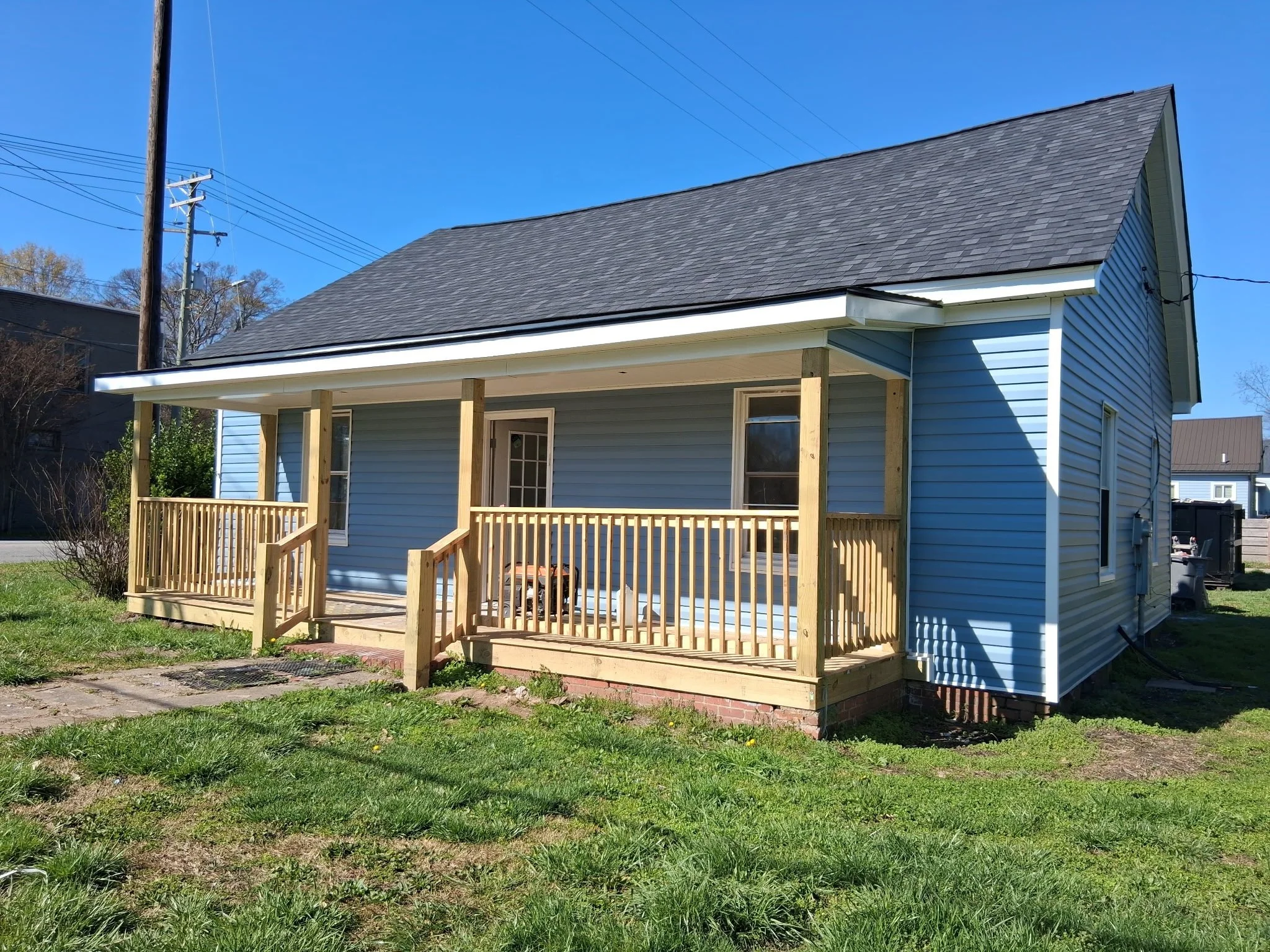 A house with blue siding and a partially built wooden porch with railings, set against a clear blue sky. There are power lines and trees in the background.
