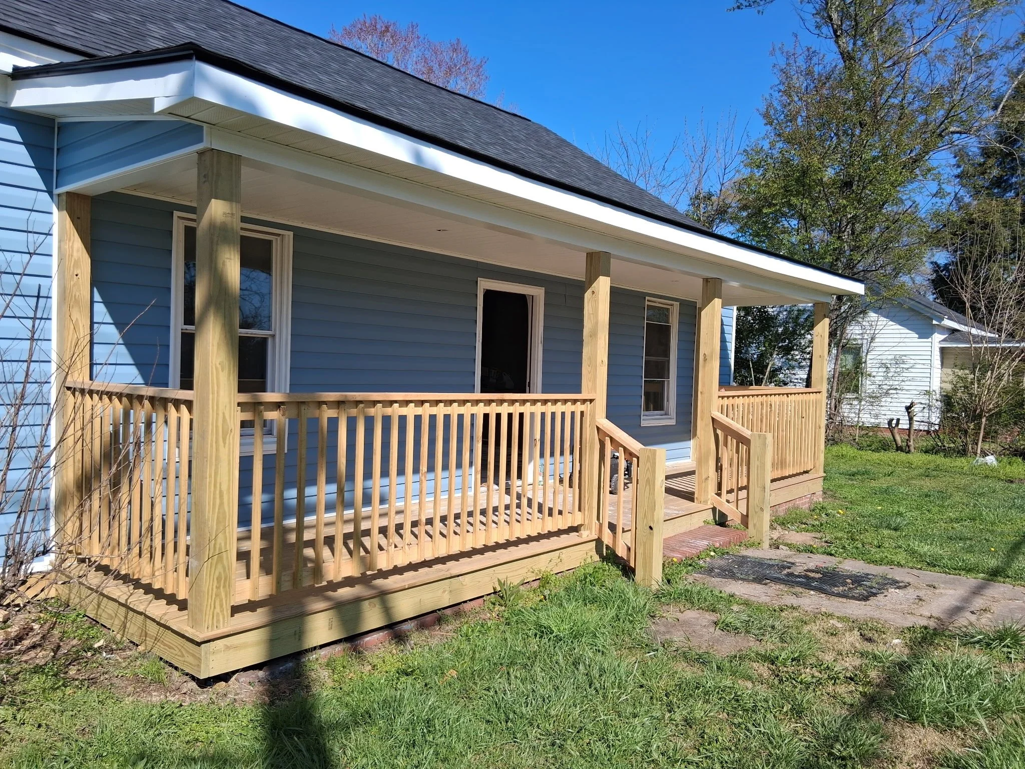 Back porch of a blue house with a wooden railing and posts, surrounded by green grass and trees, under a clear blue sky.