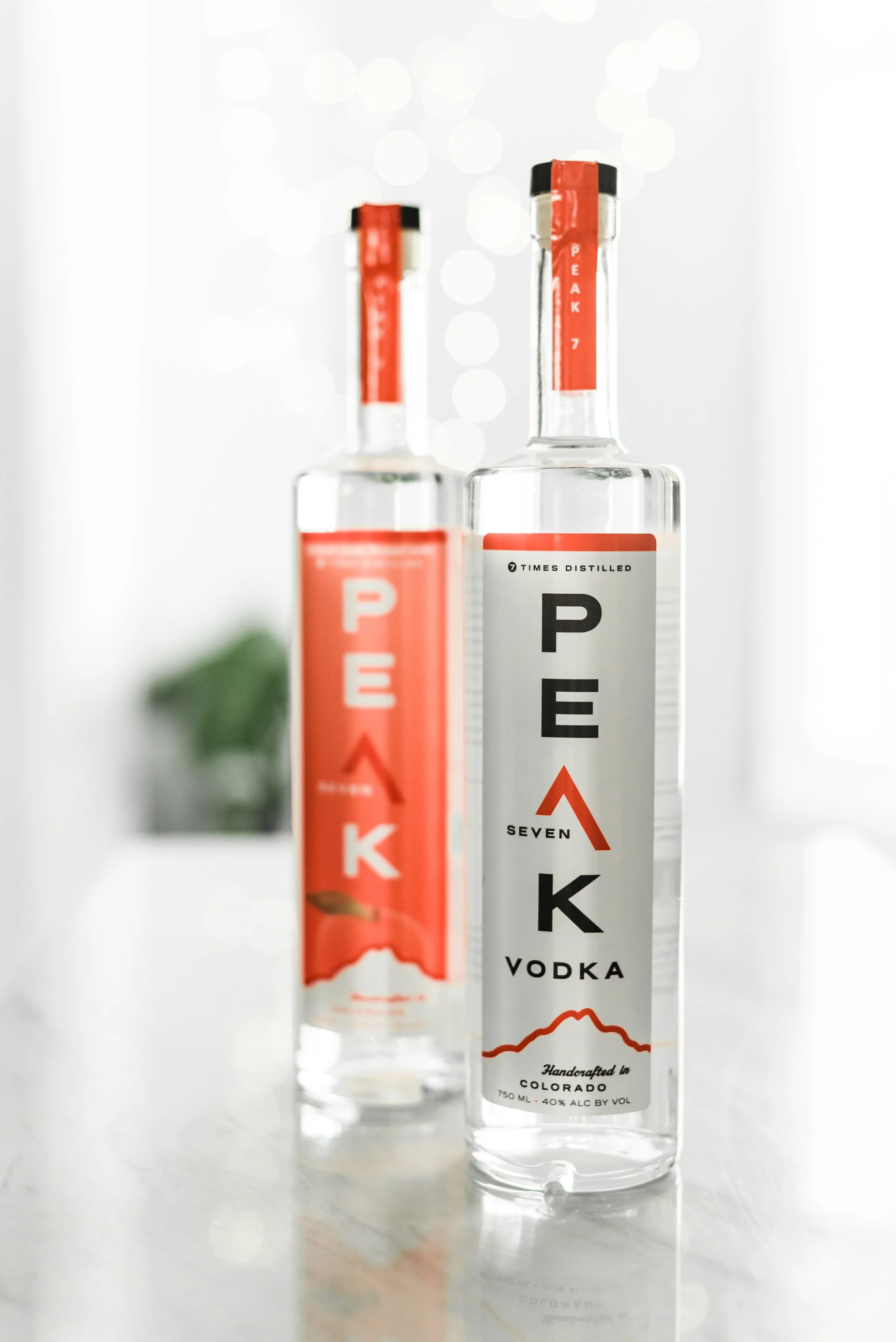 Two clear liquor bottles with white labels and red accents, one labeled 'PEAK 7 VODKA' and the other partially visible labeled 'PEAK 7 VODKA', placed on a reflective surface with bokeh lights in the background.