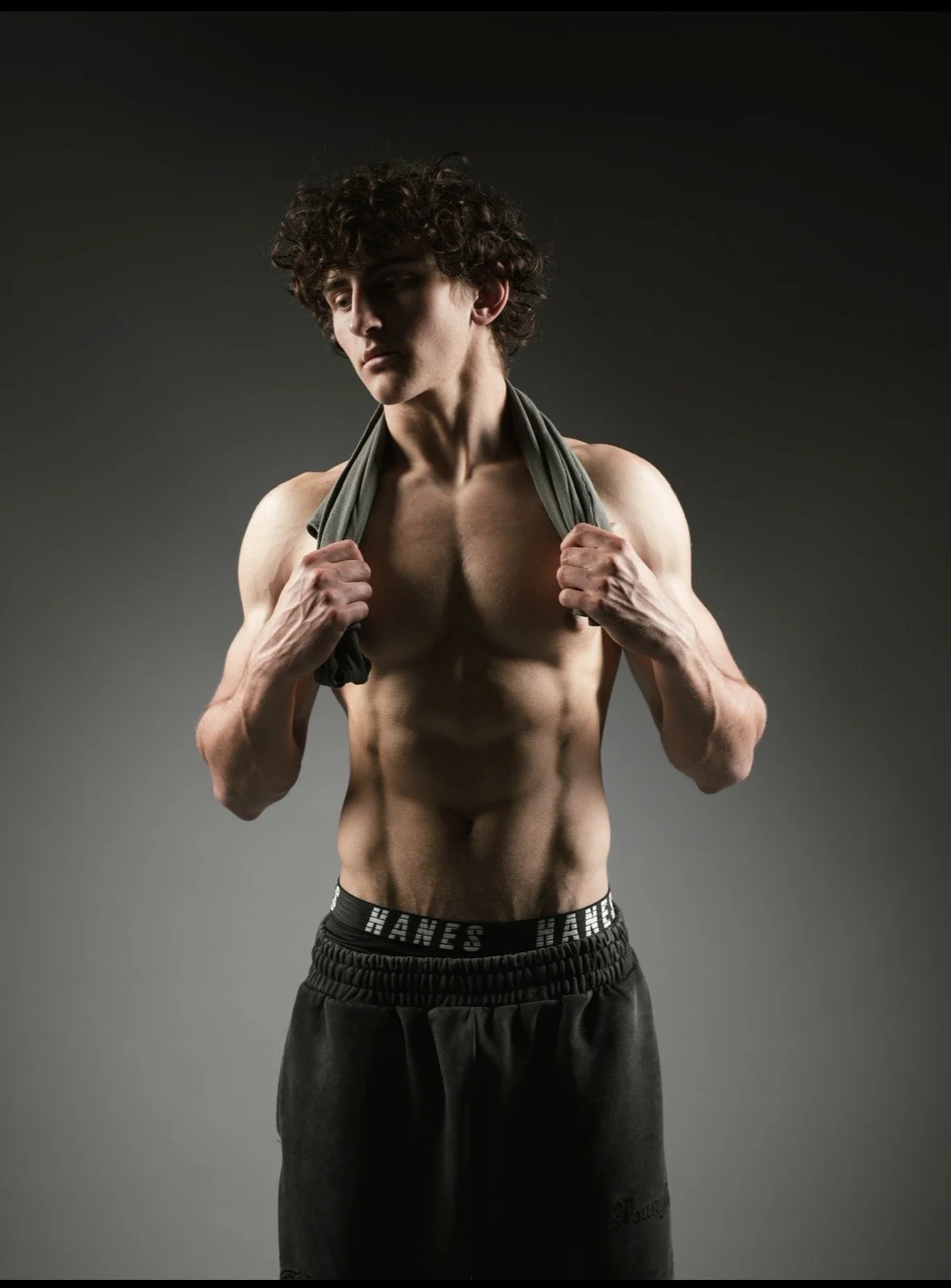 A young man with curly brown hair and a fit physique is lifting a gray shirt over his shoulders, revealing his defined abs. He is wearing black athletic pants with the brand name "Hanes" on the waistband, standing against a dark background.
