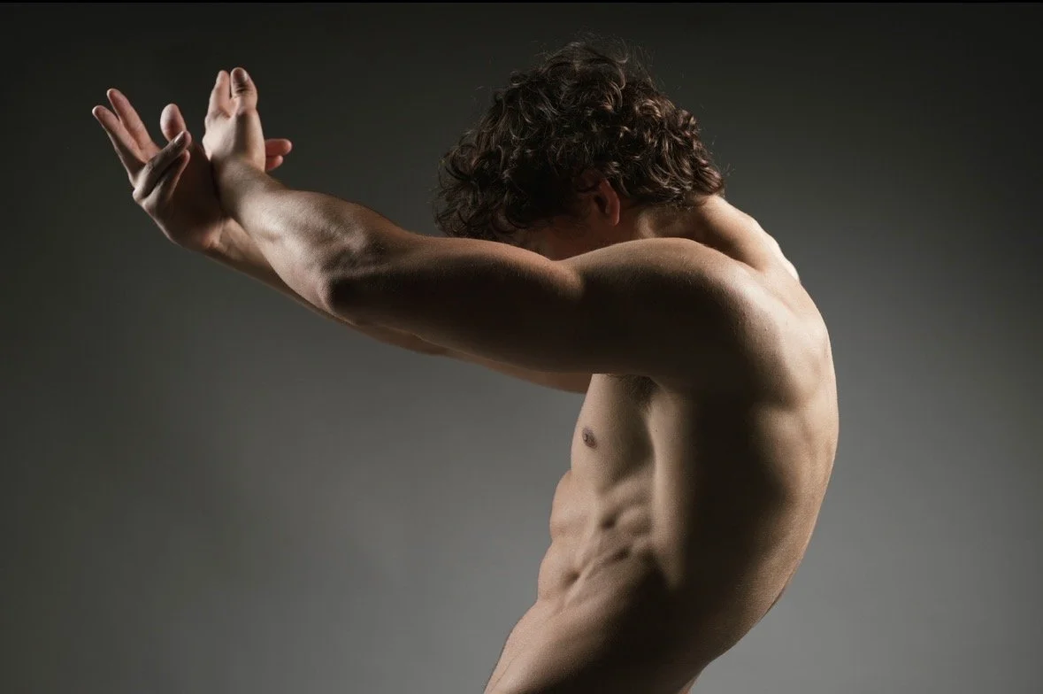 A male model with arms extended, head lowered, and body slightly curved, against a gray background.