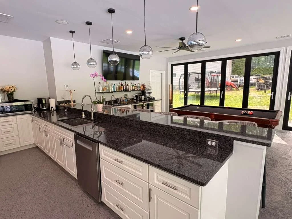 Modern kitchen with white cabinets, black granite countertops, pendant lights, and a bar area. In the background, there is a living space with a pool table and large sliding glass doors leading to a backyard with a swing set and grassy area.