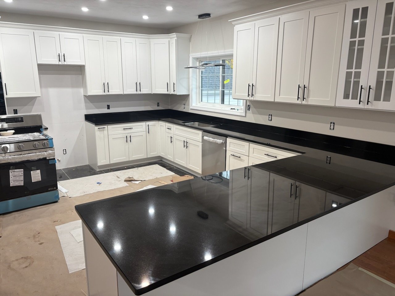Kitchen with white cabinets, black countertops, a window above the sink, and a dishwasher installed. The floor is unfinished with construction materials visible.