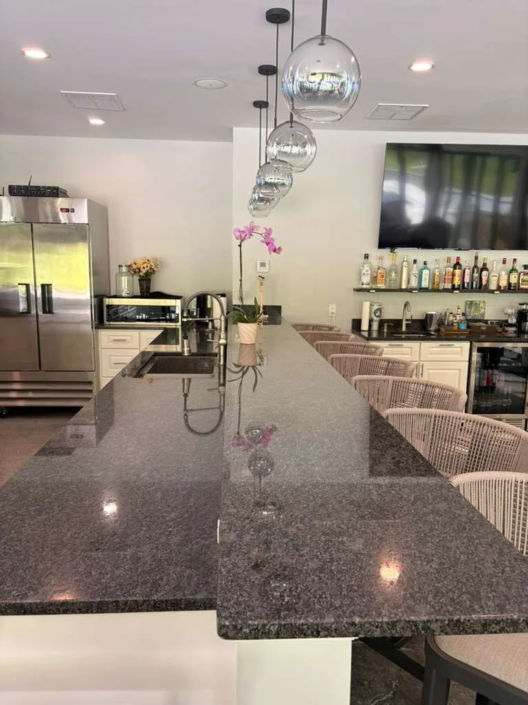 A modern kitchen with a long granite countertop, hanging spherical pendant lights, a small potted orchid, a coffee machine, a refrigerator, a flower vase, bar stools, a television, and bottles of alcohol on shelves.