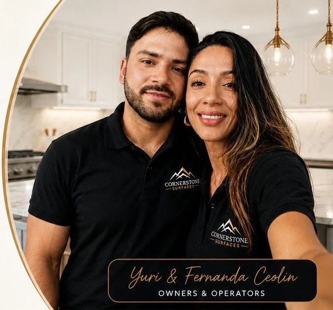 A man and woman taking a selfie in a modern kitchen, both wearing black polo shirts with the logo 'Cornerstone Surfaces.' The woman has long, dark hair with highlights and the man has dark hair and a beard. The image includes a caption with their names, 'Yuri & Fernanda Ceolin,' and titles, 'Owners & Operators.'