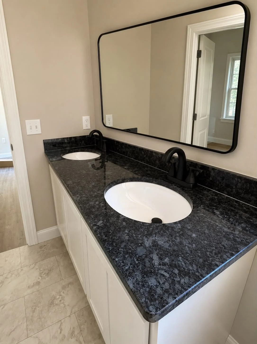 Double bathroom vanity with black granite countertop, white oval sinks, black faucets, and a large rectangular mirror above, in a neutral-colored bathroom.