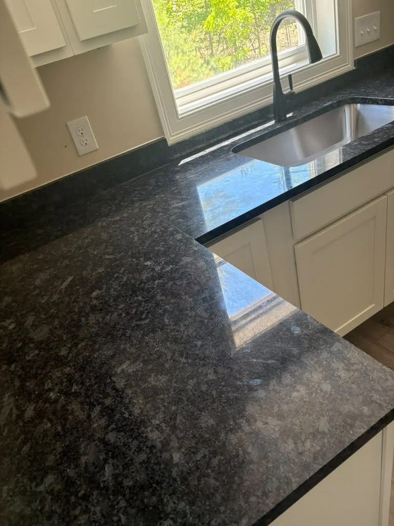 Close-up view of a black granite kitchen countertop with a built-in sink, next to a window overlooking green trees outside.