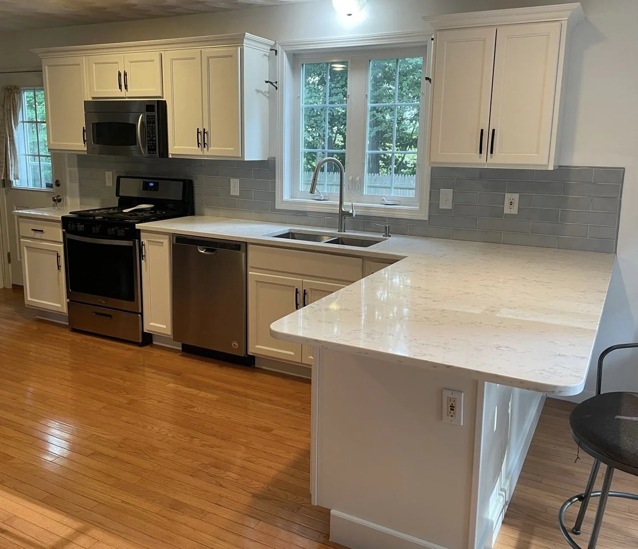 Kitchen with white cabinets, gray backsplash, marble countertop extension, wood floor, and stainless steel appliances including microwave, stove, and dishwasher. A window over the sink shows greenery outside.