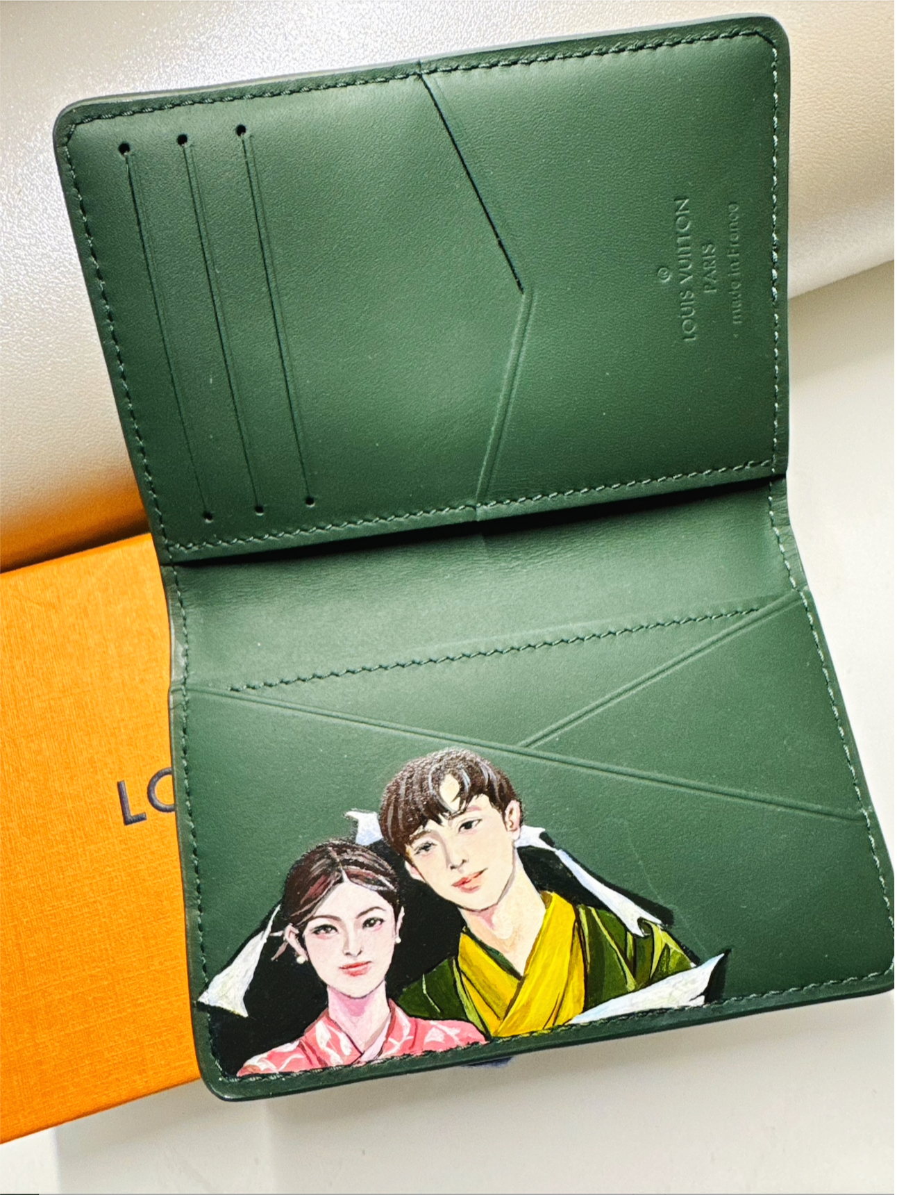Inside of a green Louis Vuitton wallet with illustrations of a young man and woman wearing traditional clothing, and an orange Louis Vuitton box underneath.