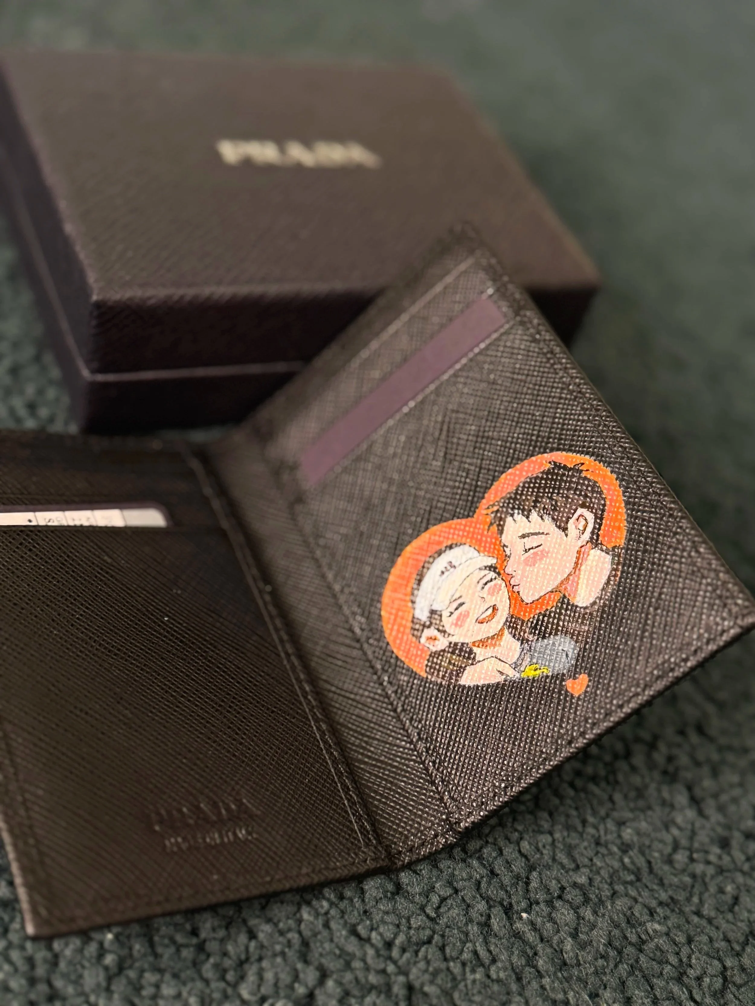A black box with a sticker of two anime-style characters hugging, with one character having orange hair. The box is on a textured dark surface.