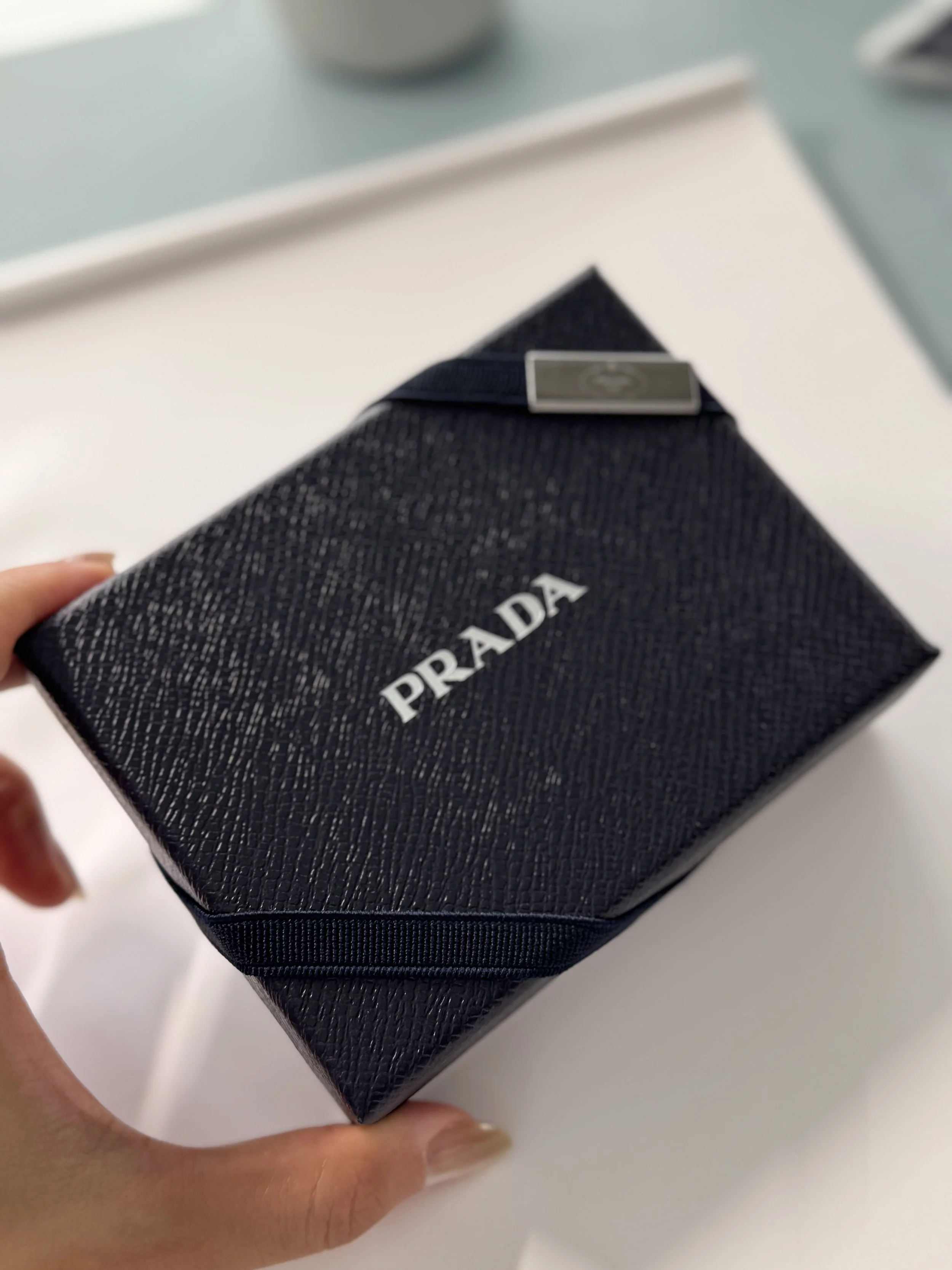 Close-up of a Prada branded black textured box being held by a person's hand.