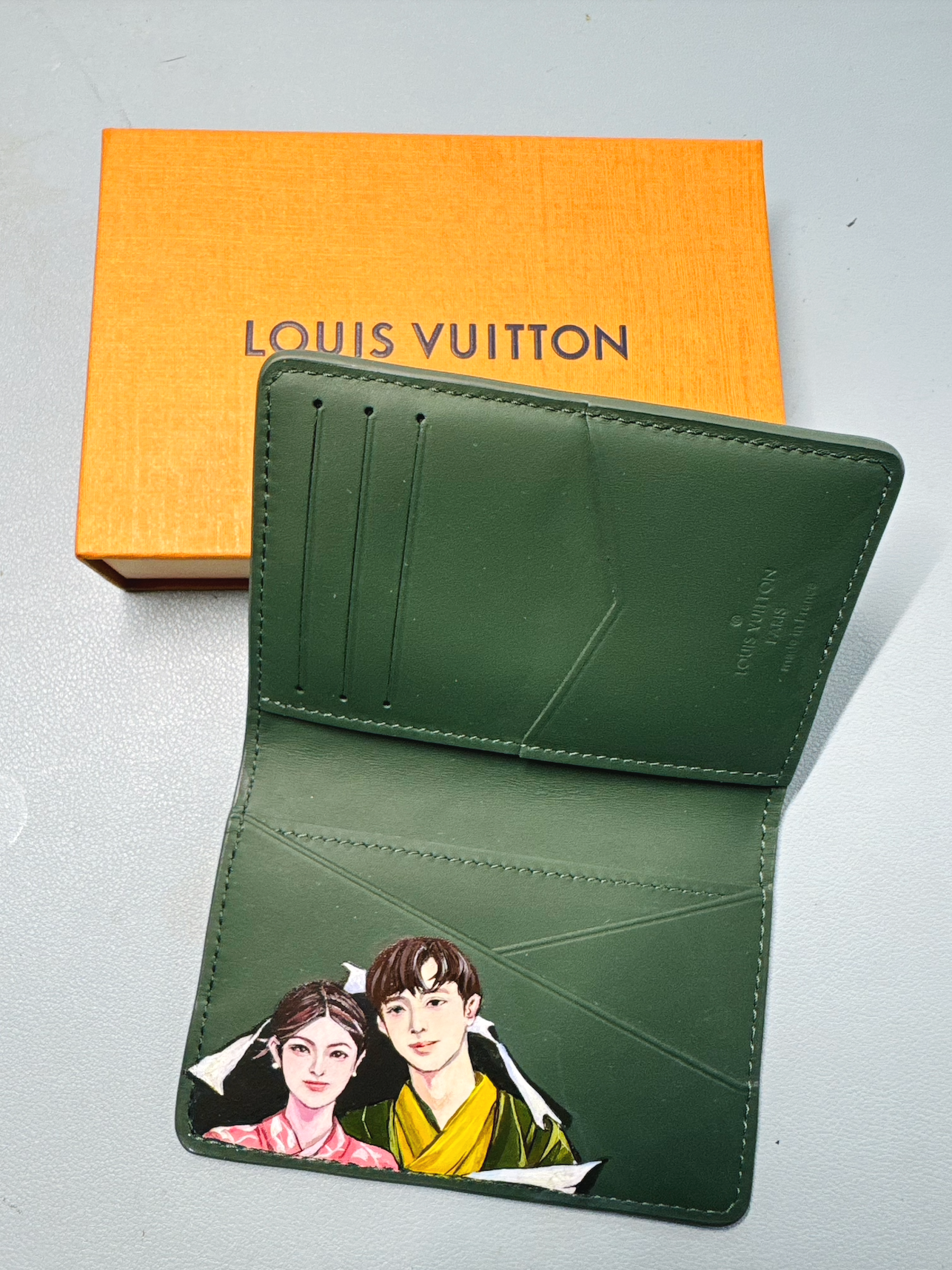 A green Louis Vuitton wallet featuring an illustration of a man and woman dressed in traditional Asian clothing, with an orange Louis Vuitton box in the background.