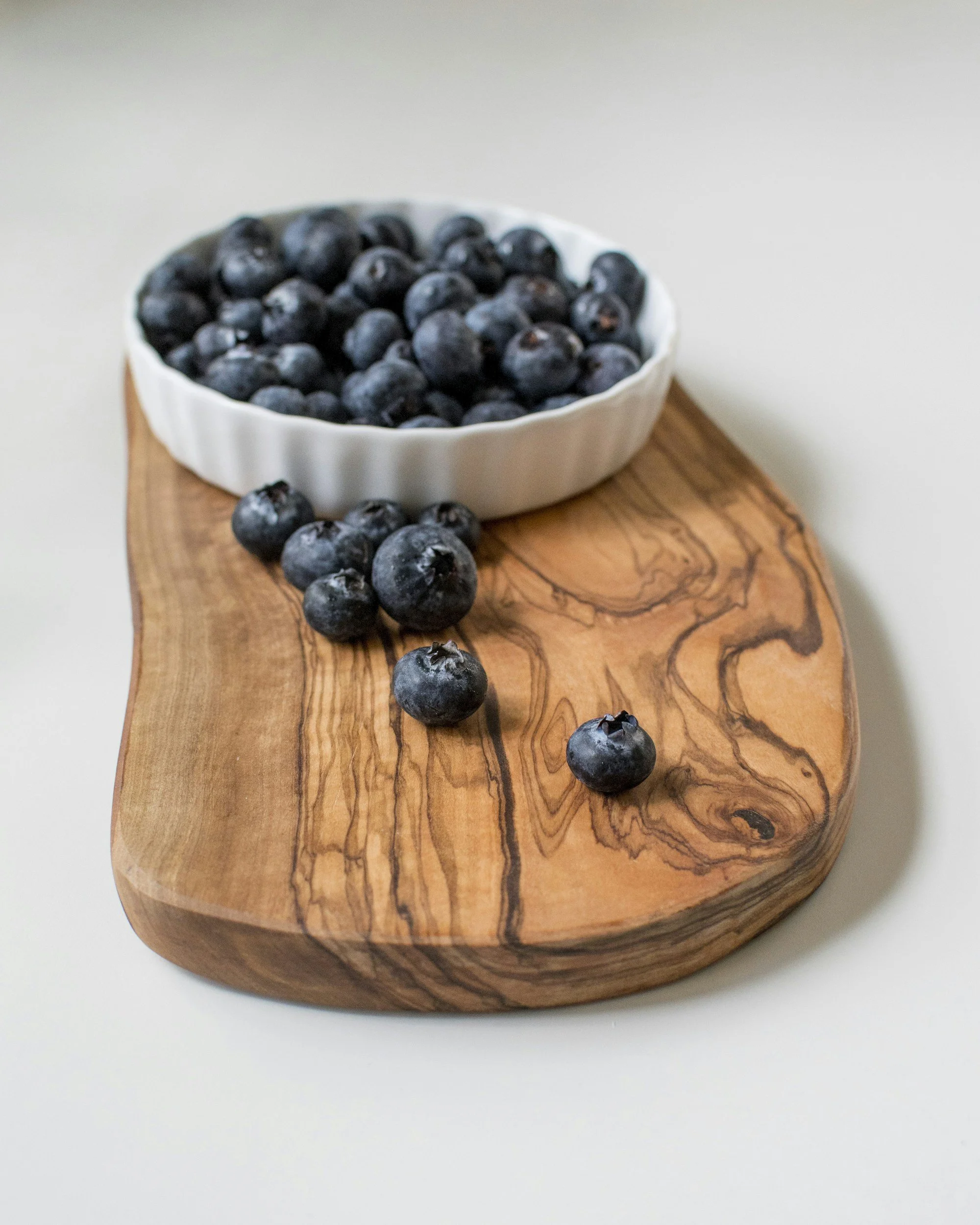 A white bowl filled with blueberries on a wooden cutting board, with some blueberries spilled on the board, against a plain light-colored background.