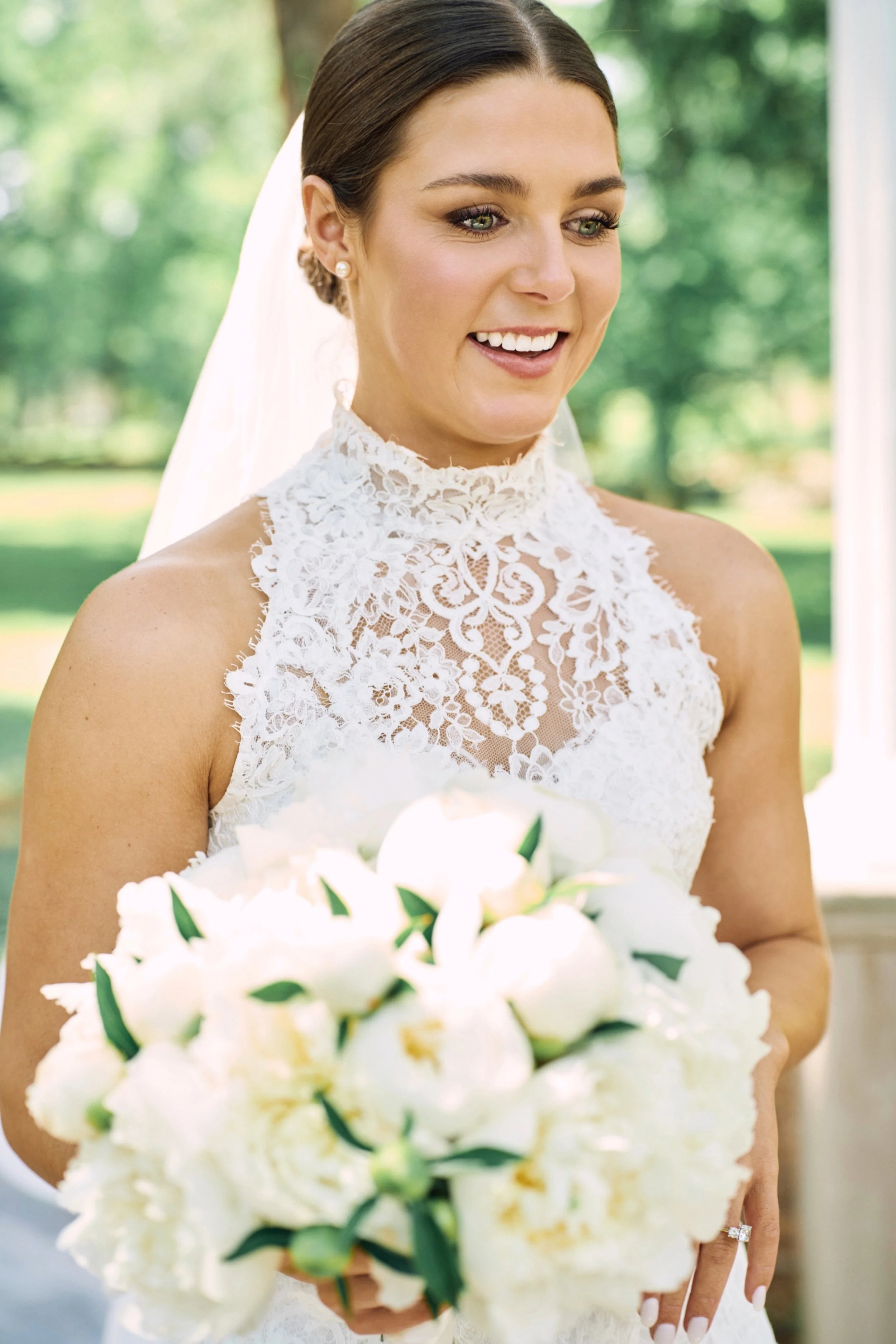 A bride with dark brown hair styled in an updo wearing pearl earrings and a white lace wedding dress, holding a bouquet of white flowers with green leaves, outside on a sunny day.