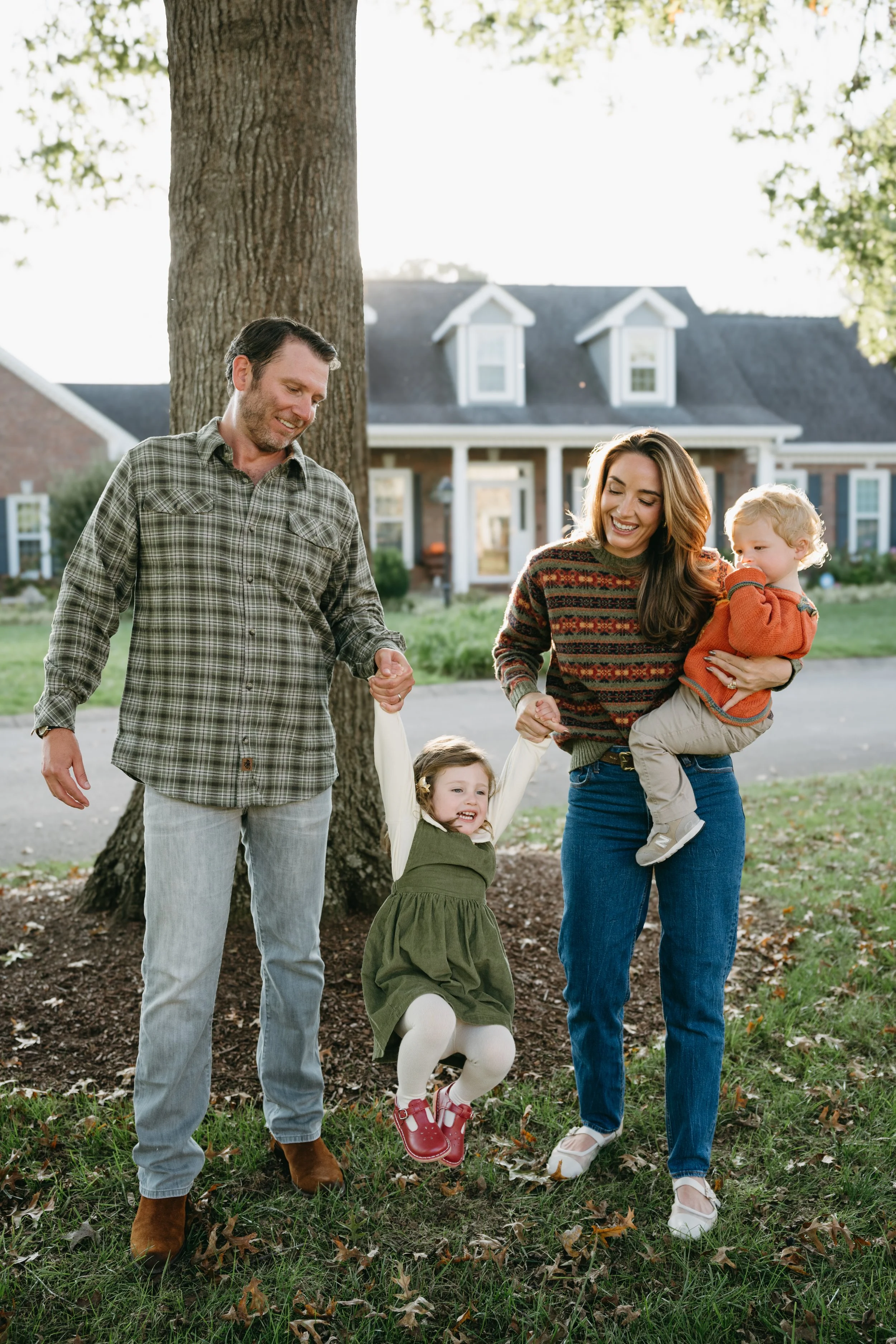A family with two children enjoying a walk outdoors in front of a house with white trim, large trees, and green grass, during daylight.