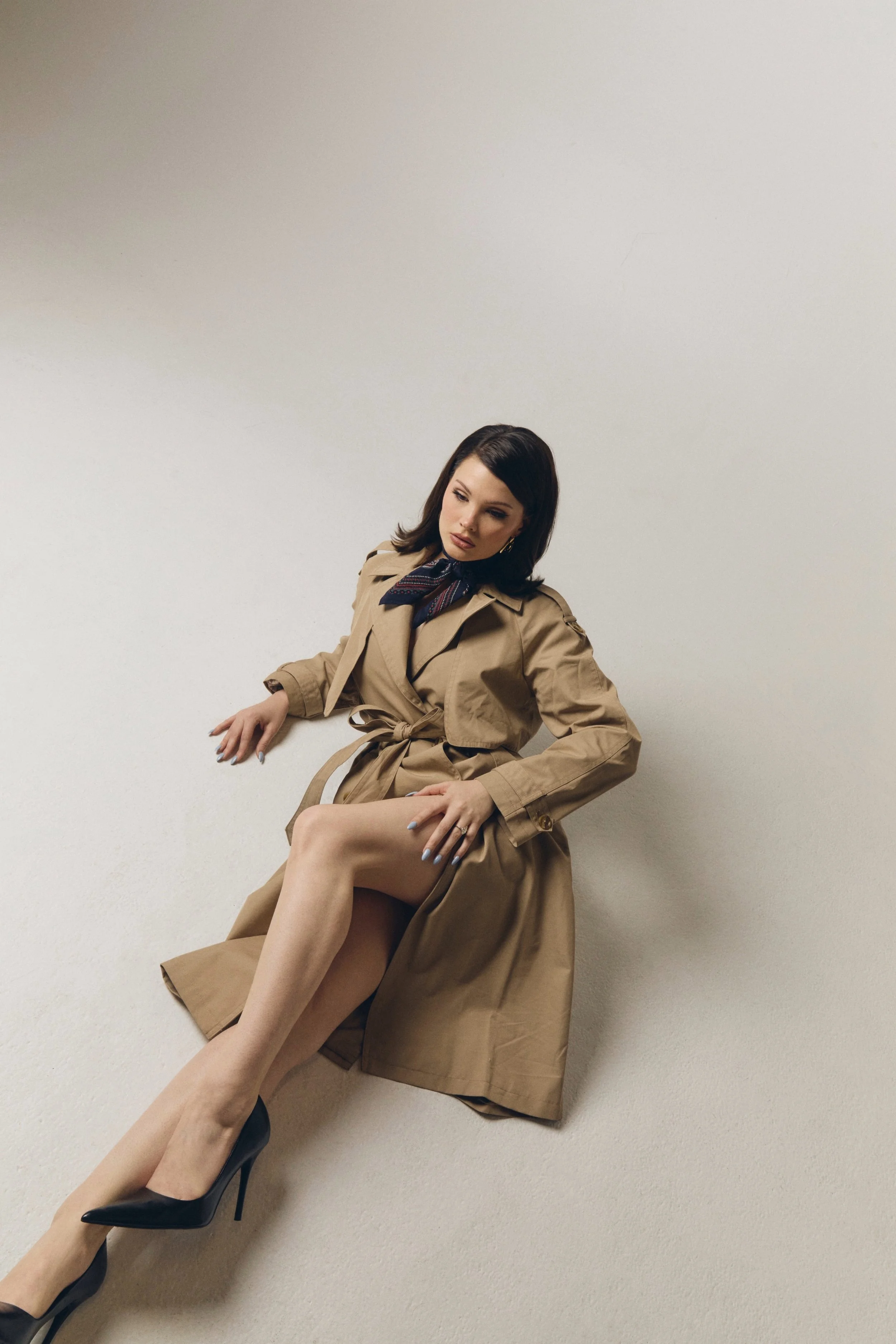 A woman with dark hair, wearing a beige trench coat, sitting against a plain white wall, with her legs crossed and wearing high heels.