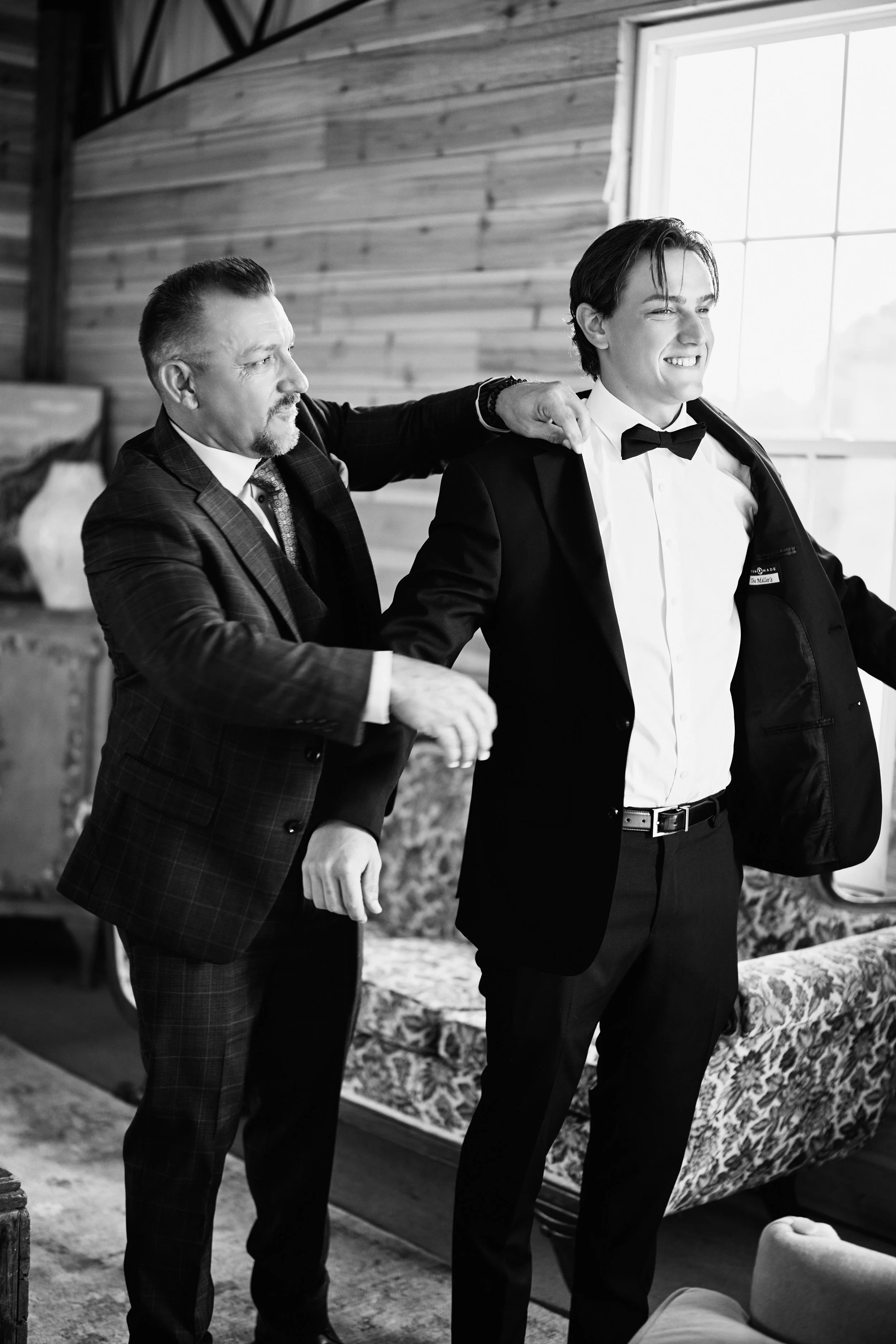 A man with slicked-back hair and a beard wearing a checkered suit and tie is helping a young man in a tuxedo with a bowtie put on a jacket in a warmly-lit room with wooden walls.