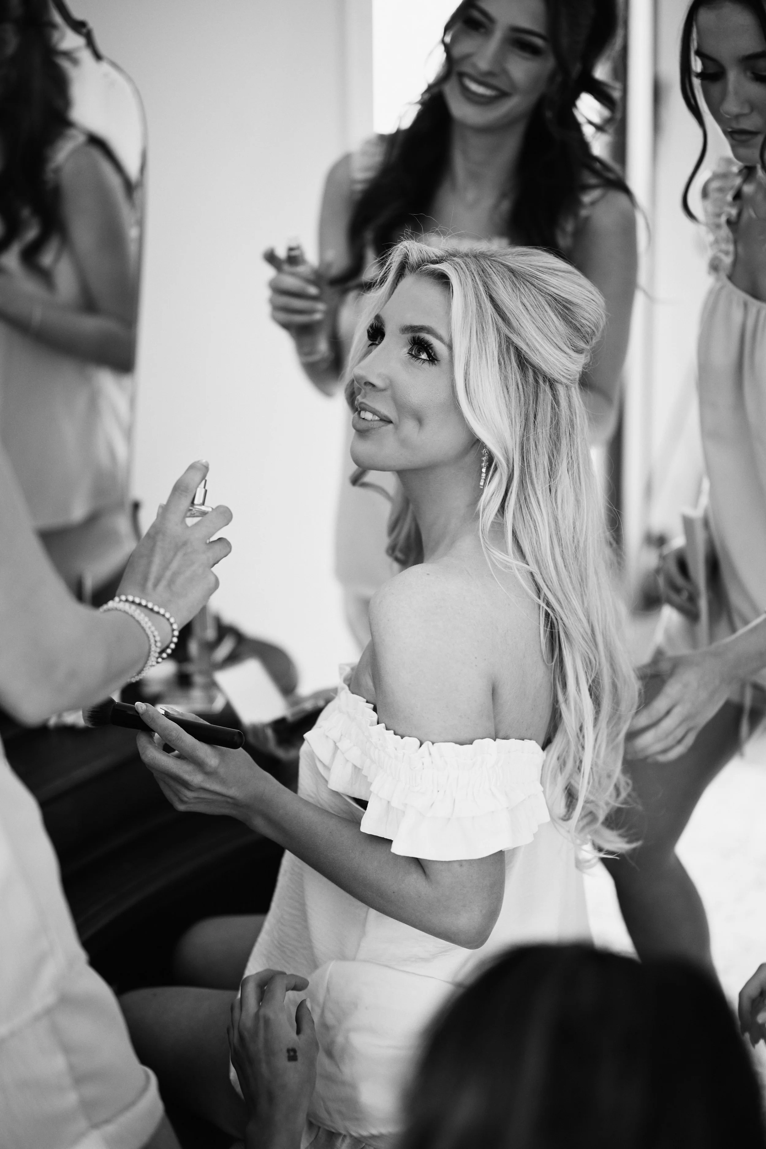 A woman with long blonde hair and an off-the-shoulder white dress is sitting, smiling and looking up as a makeup artist applies makeup to her face. Several women with dark hair, wearing dresses, are gathered around in the background, preparing for a 
