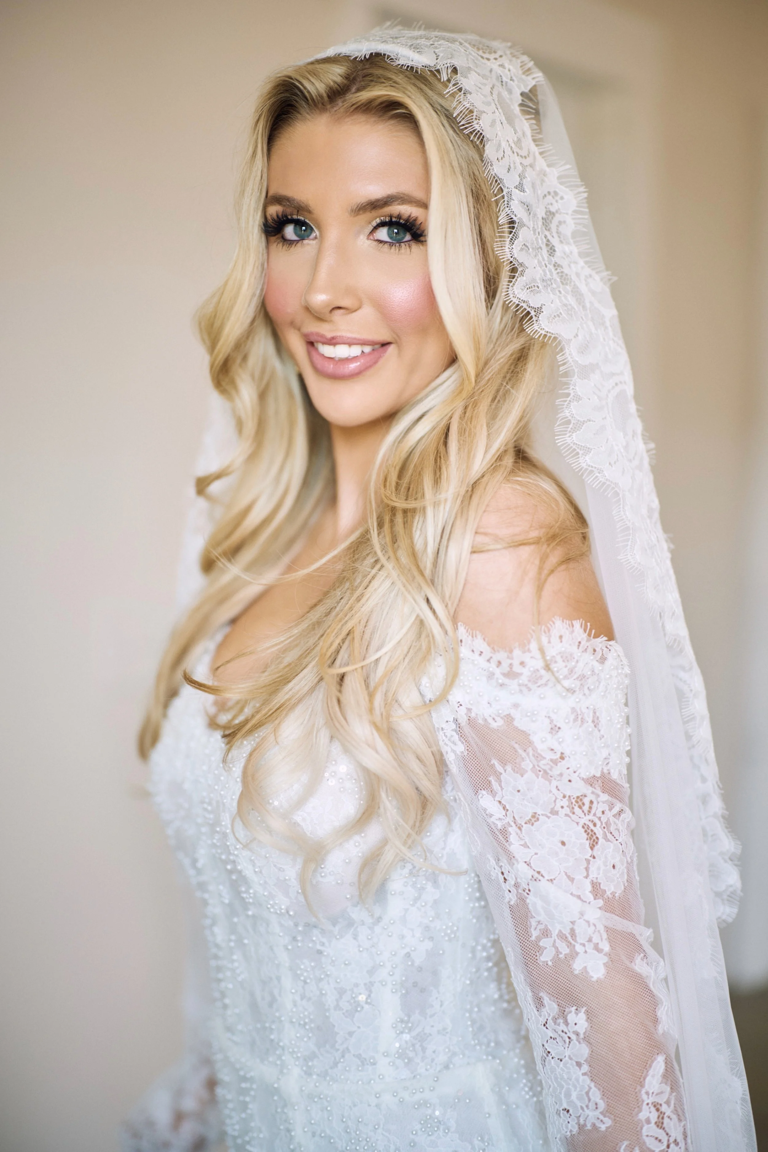 Close-up of a woman with long blonde hair wearing a white lace bridal gown and veil, smiling softly, against a neutral background.