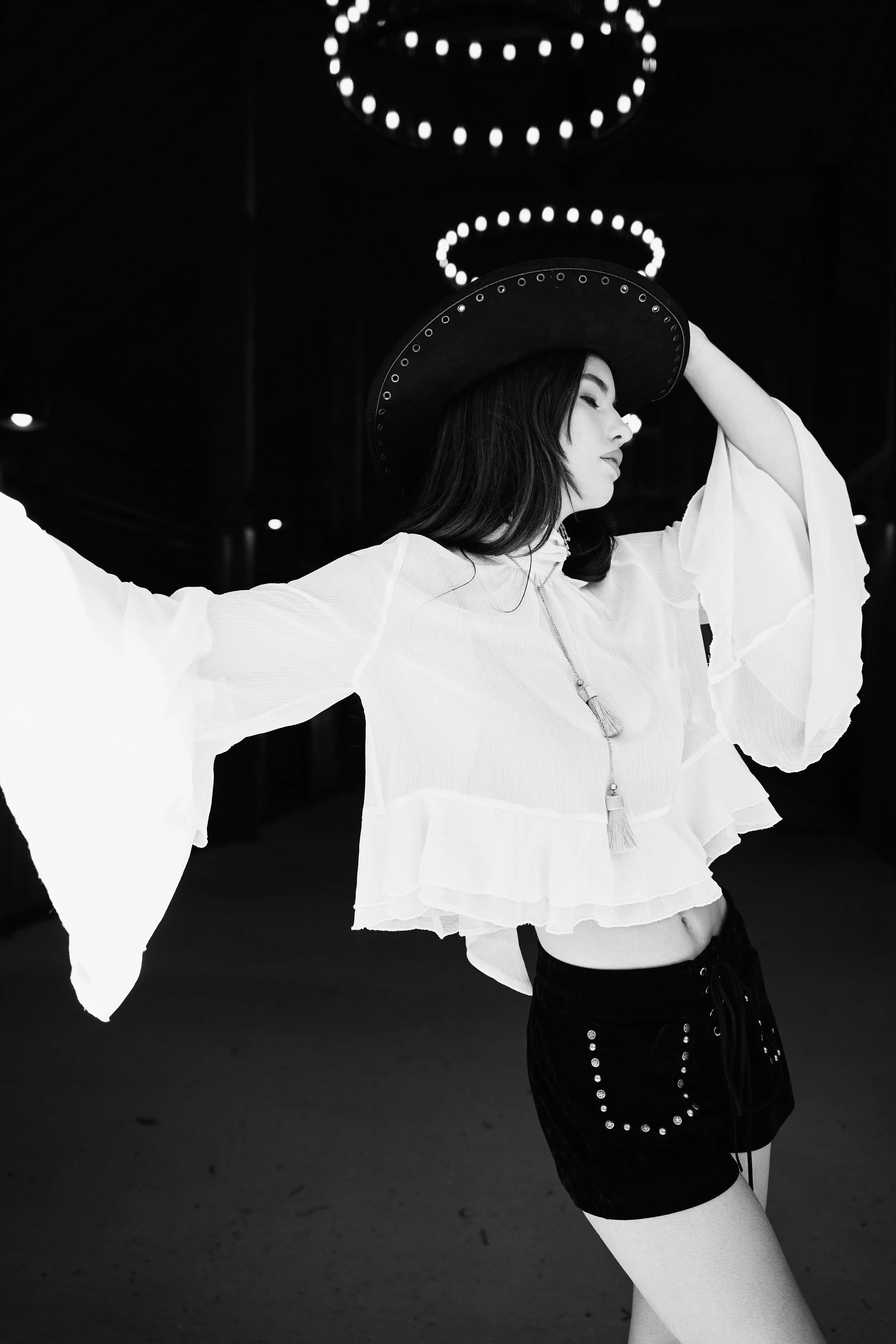 A woman in a wide-brimmed hat with metal eyelets, white flowing blouse with bell sleeves, black shorts with lace-up sides and decorative studs, and thigh-high fishnet stockings, poses with her eyes closed and one arm raised, in front of a dark backgr